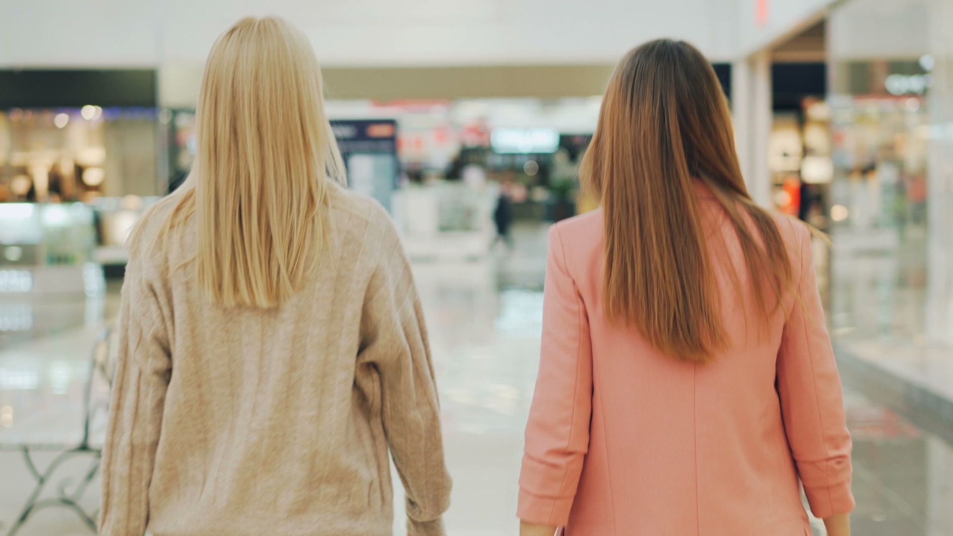 Two women walking in a mall holding shopping bags, back view.