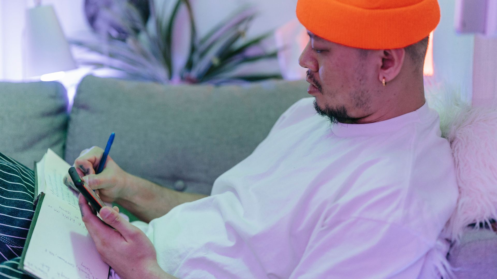 Asian man in orange hat writing in notebook while on phone. Relaxed indoor setting.