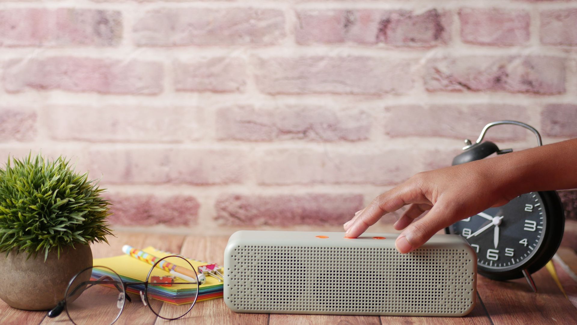 Close-up of a hand touching a wireless speaker on a desk with various office items.