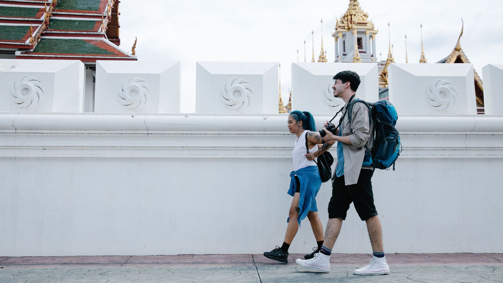 A couple with backpacks walks past stunning Thai temple architecture in an urban setting.