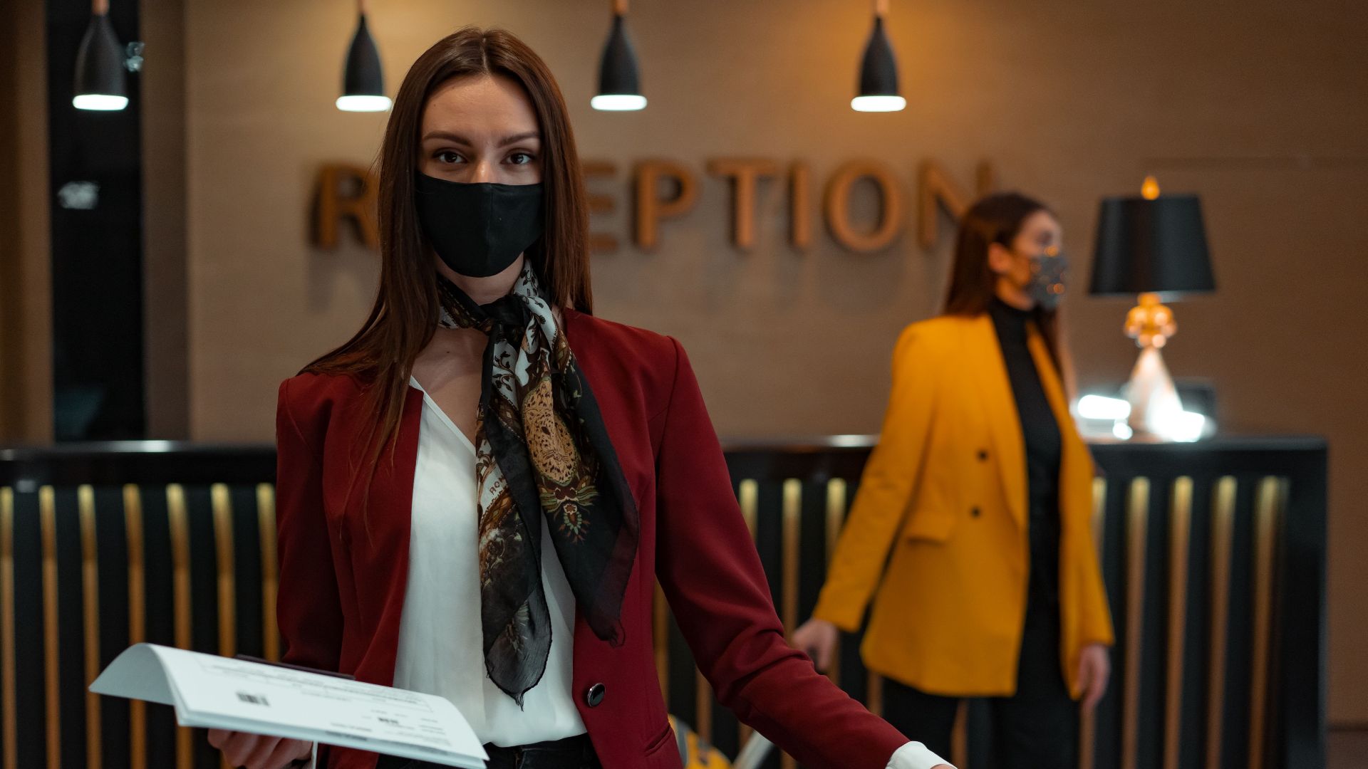 A woman in a mask stands at a hotel reception desk holding paper, welcoming service