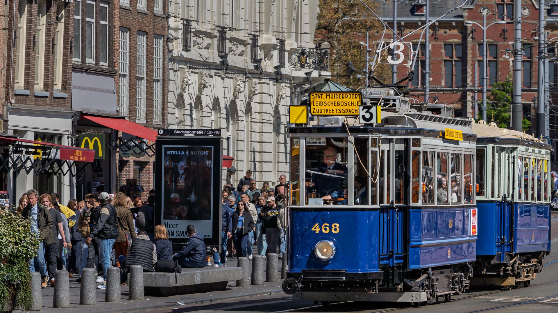 A historic blue tram rides through Amsterdam's busy street, capturing city life and classic architecture.
