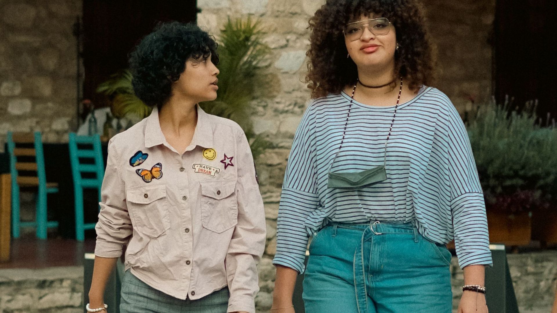 Two women with curly hair walking on stone pavement, showcasing casual fashion.