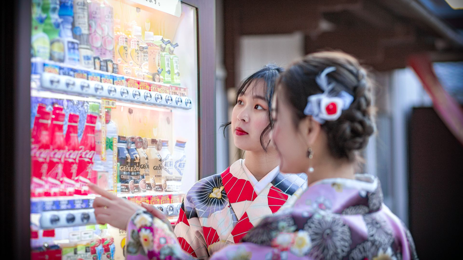 Two women wearing kimonos choose drinks from an outdoor vending machine during the day.