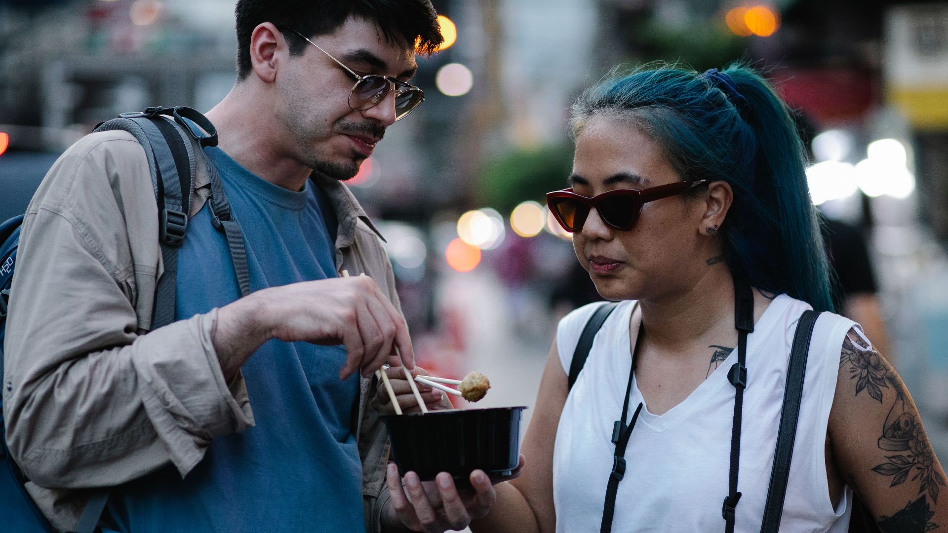 Two travelers savoring street food at night in a bustling urban setting.