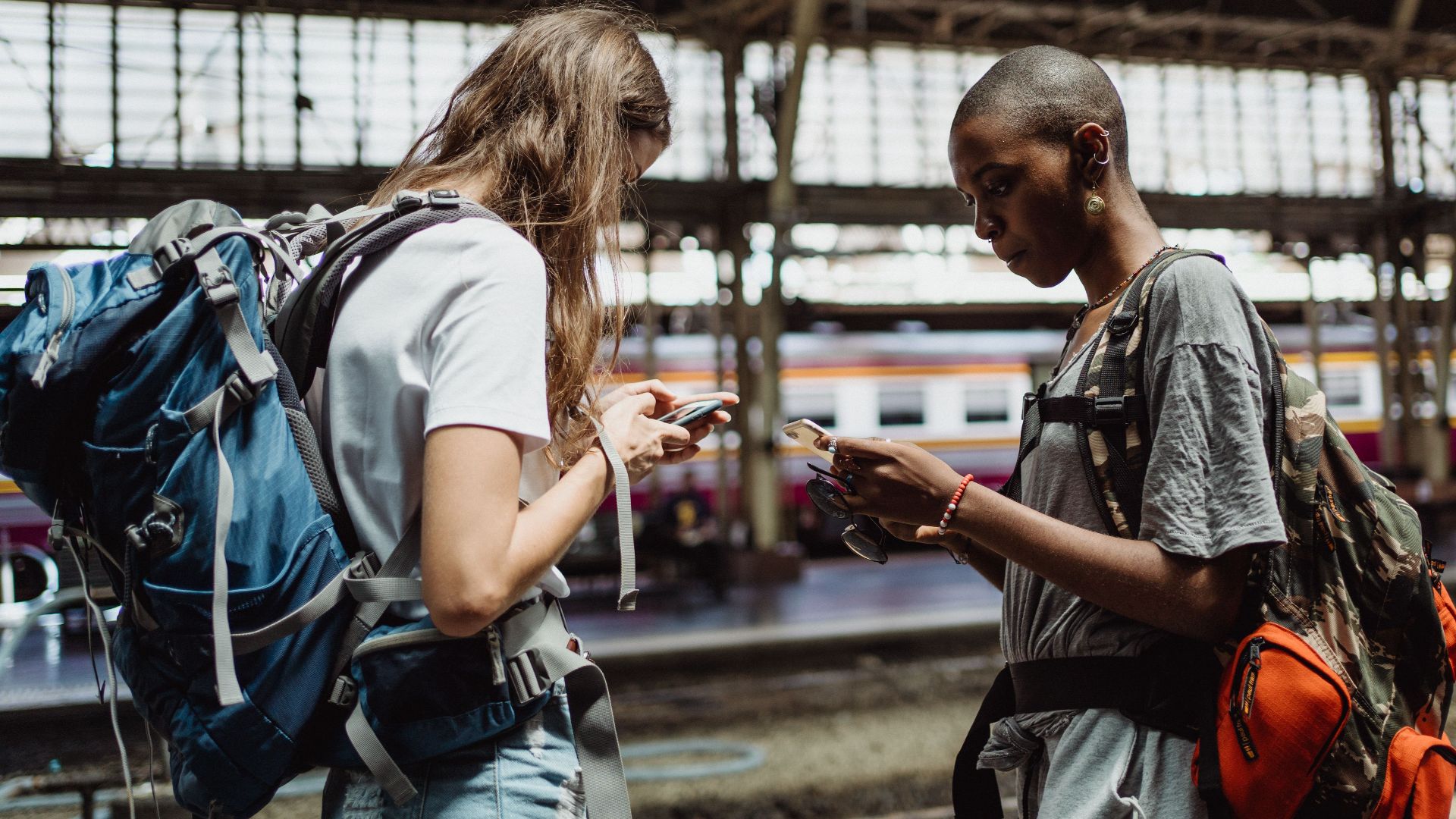 Two adventurous backpackers using smartphones while waiting at a bustling train station.