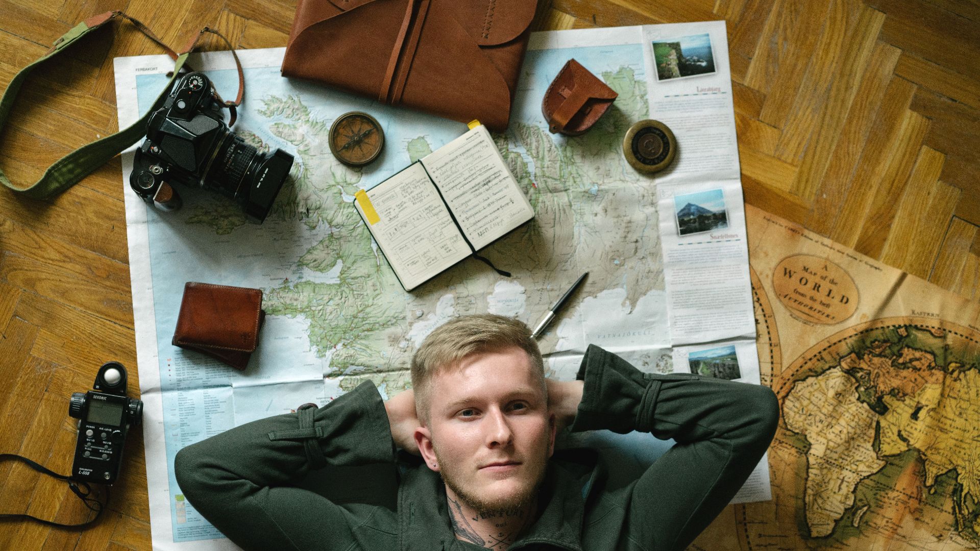 Young man dreams of travel, surrounded by maps, camera, and notebook.