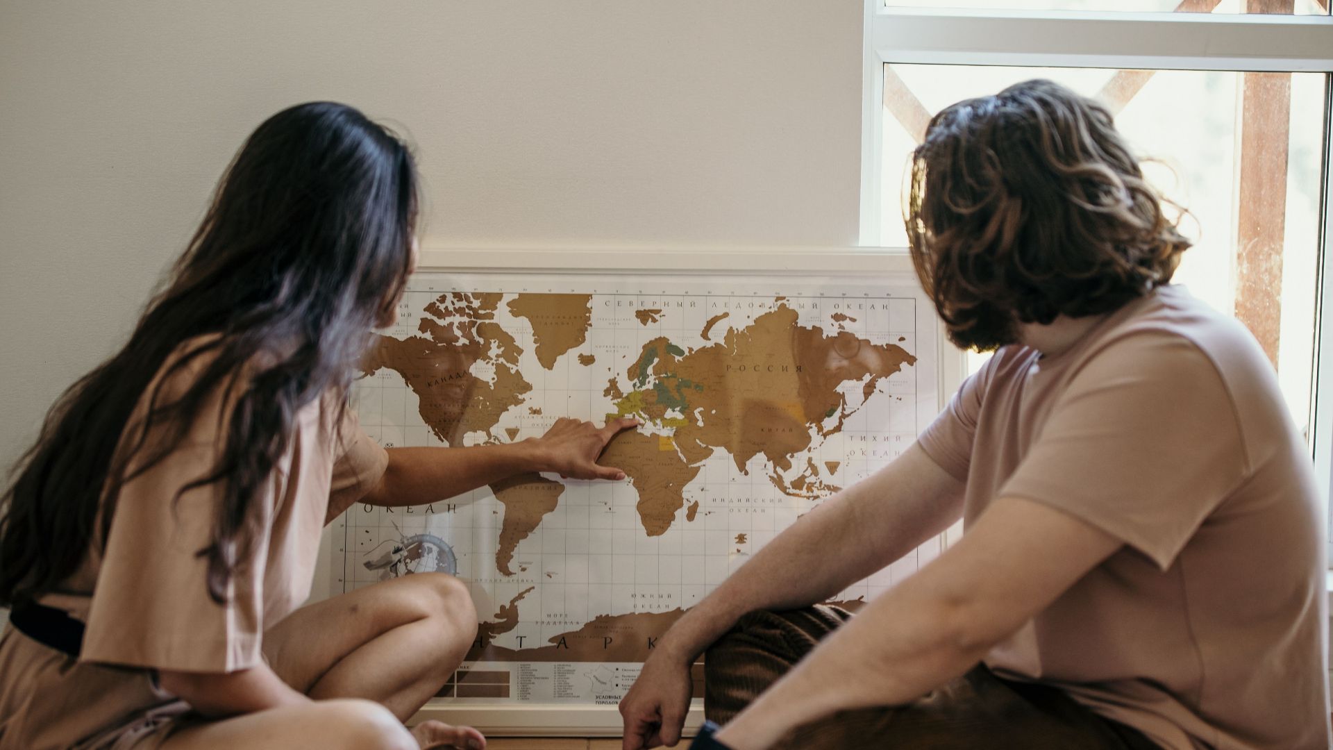 Couple sitting on floor pointing at a world map, planning their next travel adventure indoors.