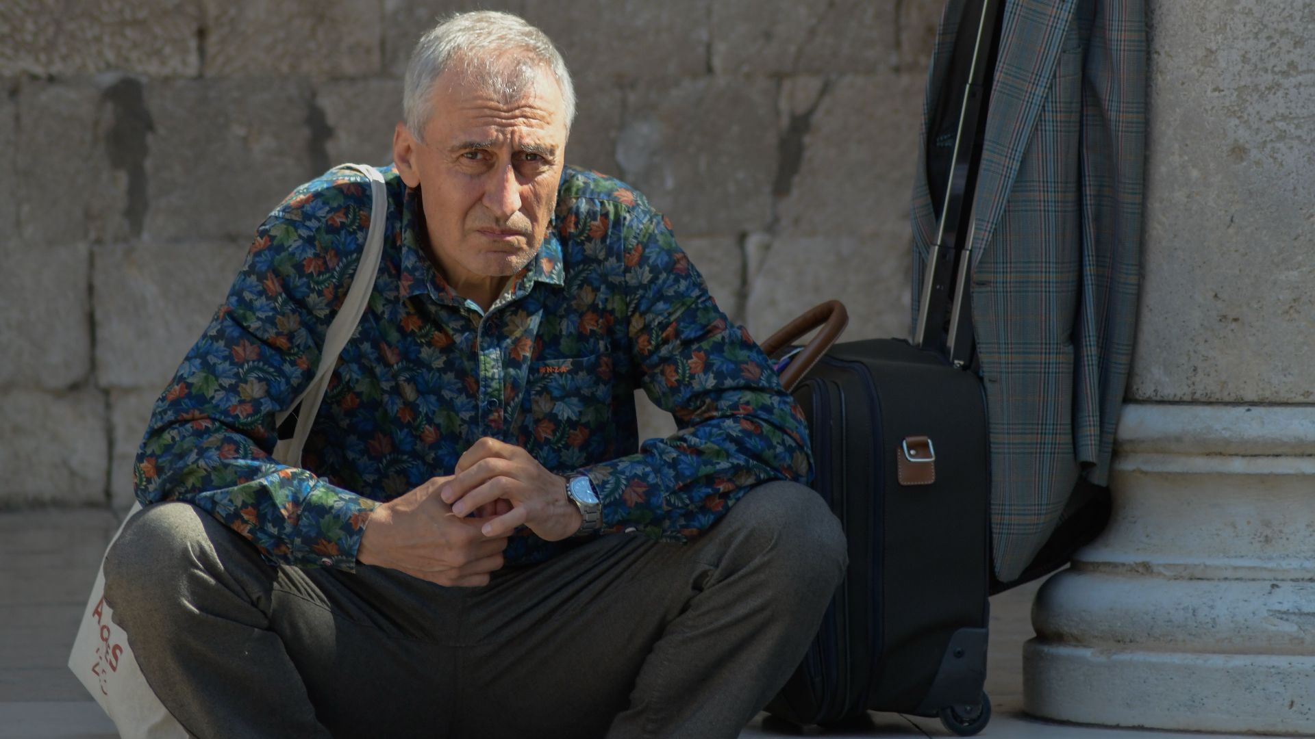 Senior tourist with luggage sits thoughtfully in historic Dubrovnik location.