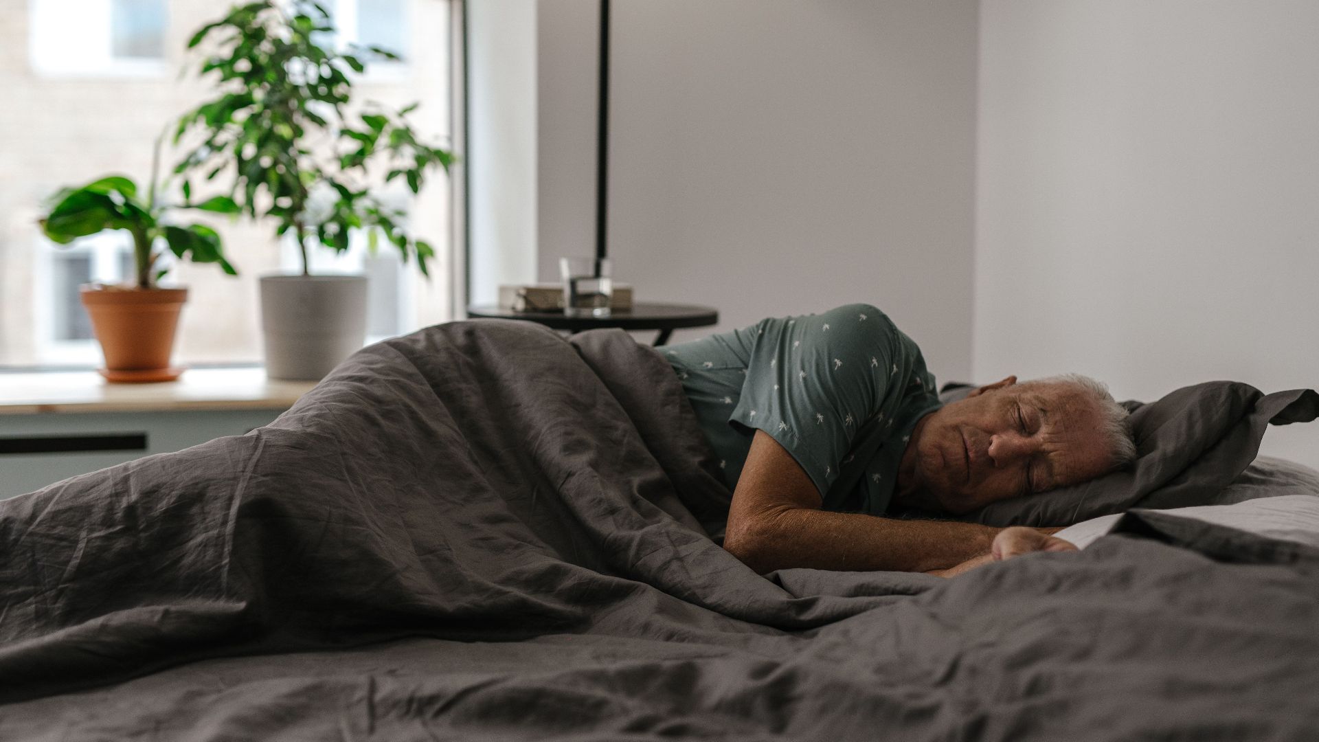 Senior man resting peacefully on a bed inside a room with natural light and plants.