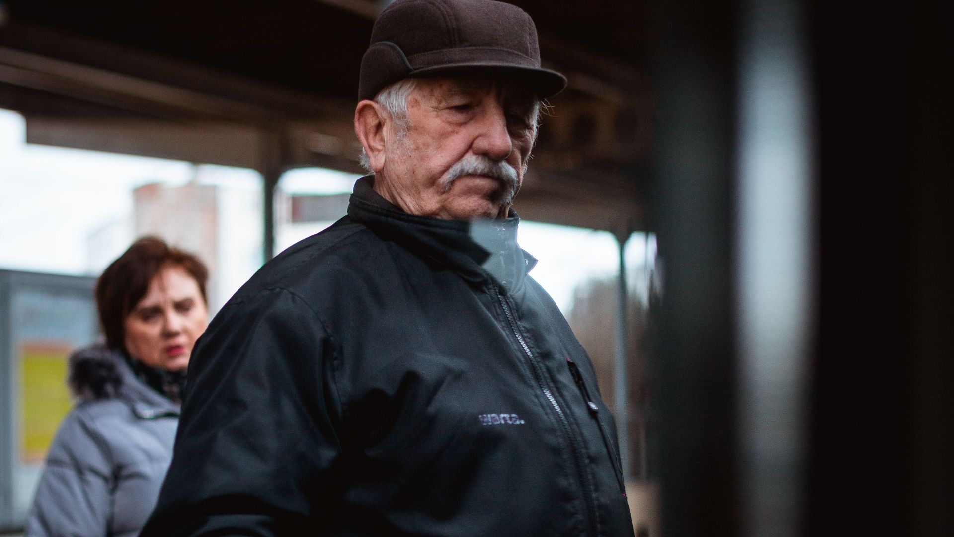 Senior man in a cap and jacket standing at a train station platform.
