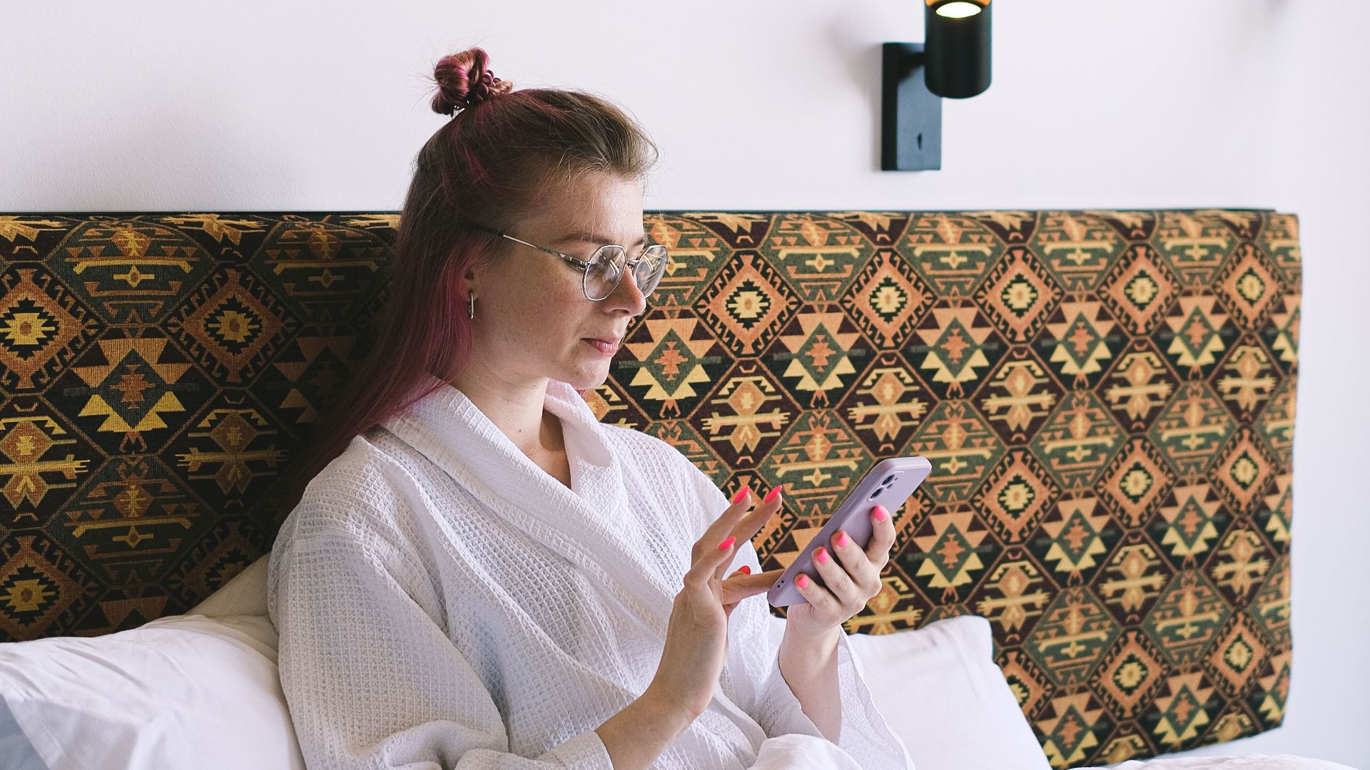 Woman in white robe using a smartphone while relaxing on a hotel bed with modern decor in the background.