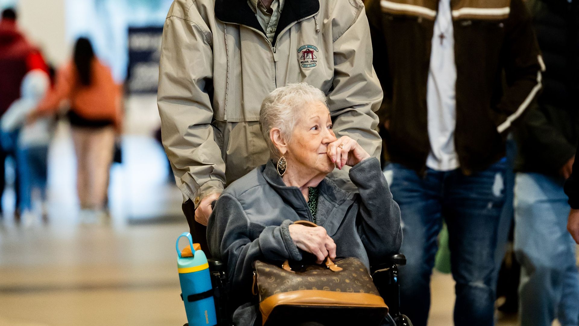 Senior woman in a wheelchair being assisted by a man in a bustling shopping mall.