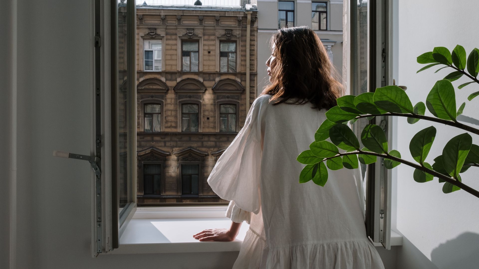 Woman in a white dress looking out an open window at classic architecture on a sunny day.