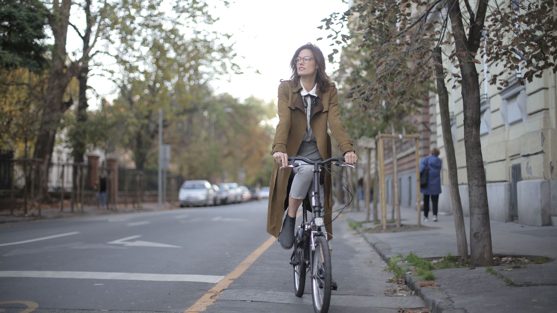 A woman enjoys cycling through a city street lined with autumn trees, embracing a leisurely lifestyle.