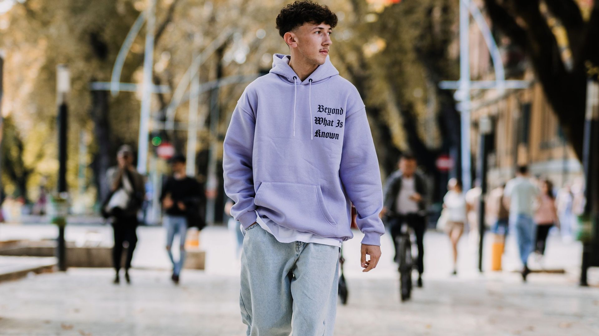 A teenager wearing a hoodie casually walks down a tree-lined street in a German city.