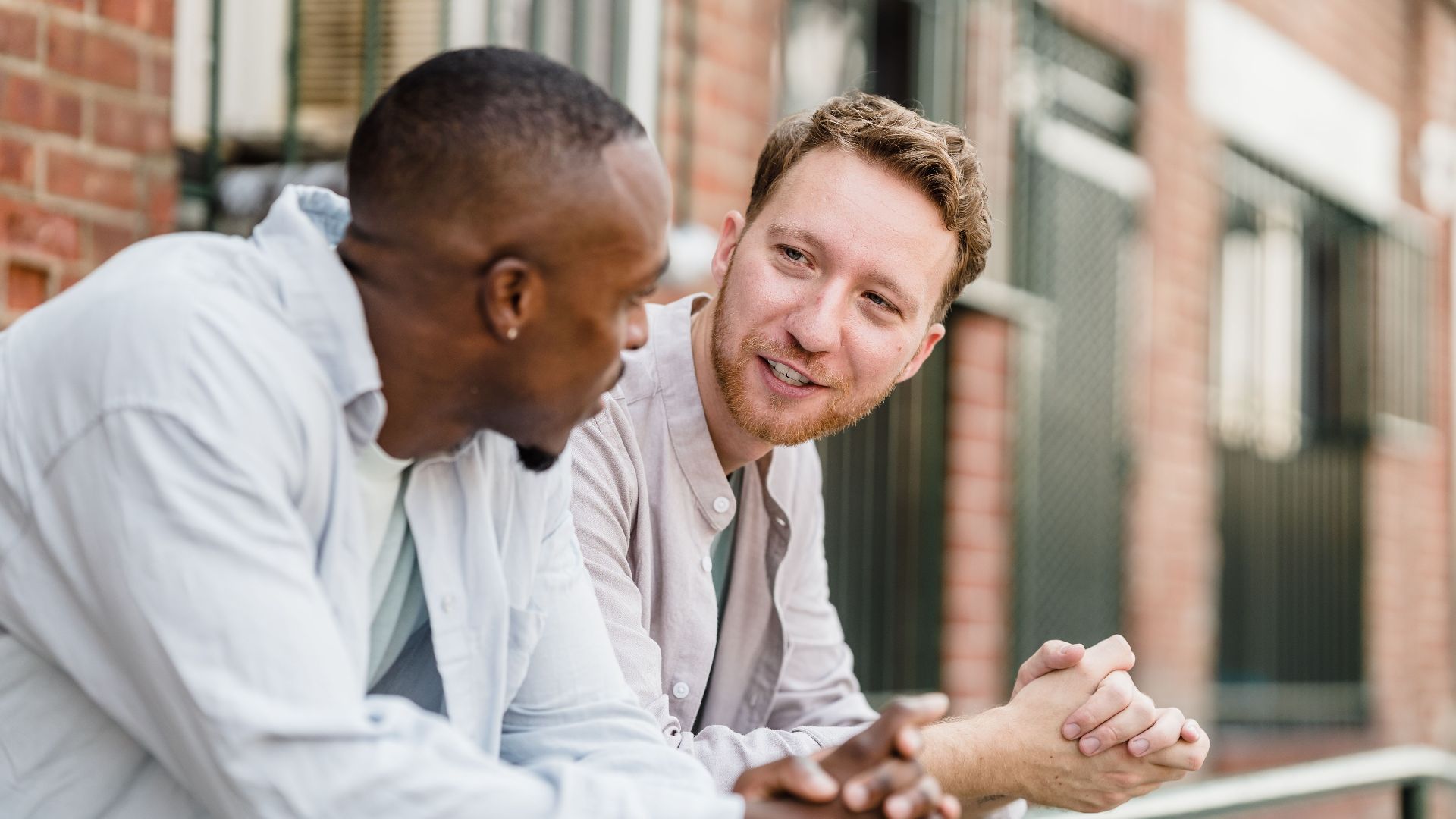 Two men engaging in lively conversation outdoors against a brick wall background.