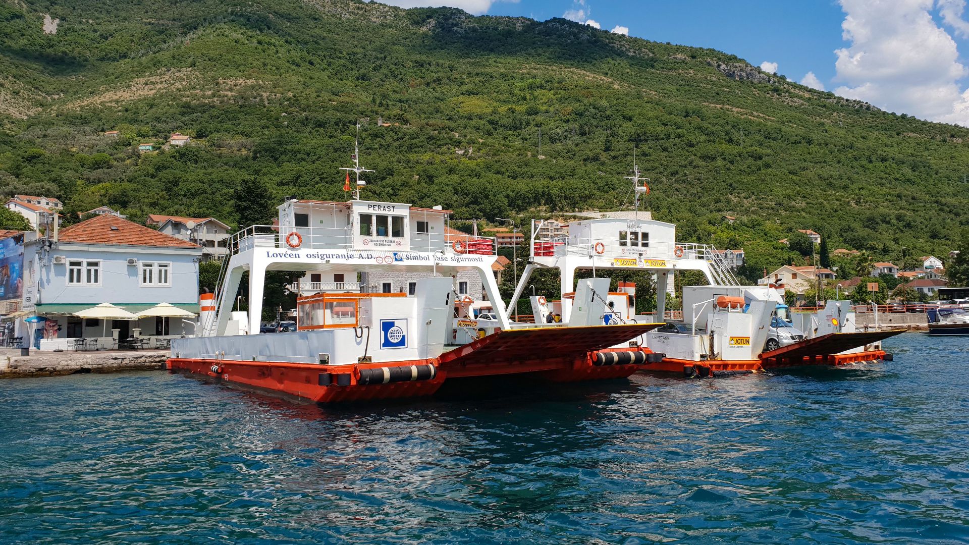 Ferries docked by lush mountains in Boka Kotorska Bay under a bright blue sky.