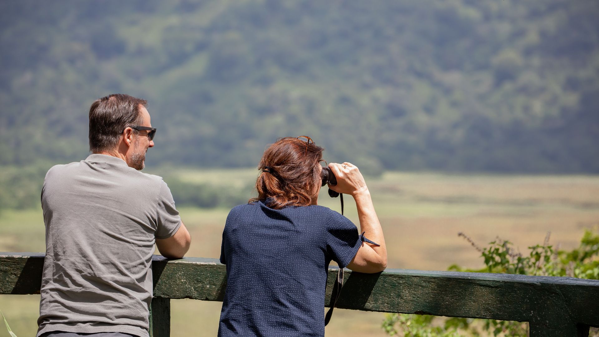 A couple relaxing outdoors, using binoculars to enjoy a scenic landscape view.