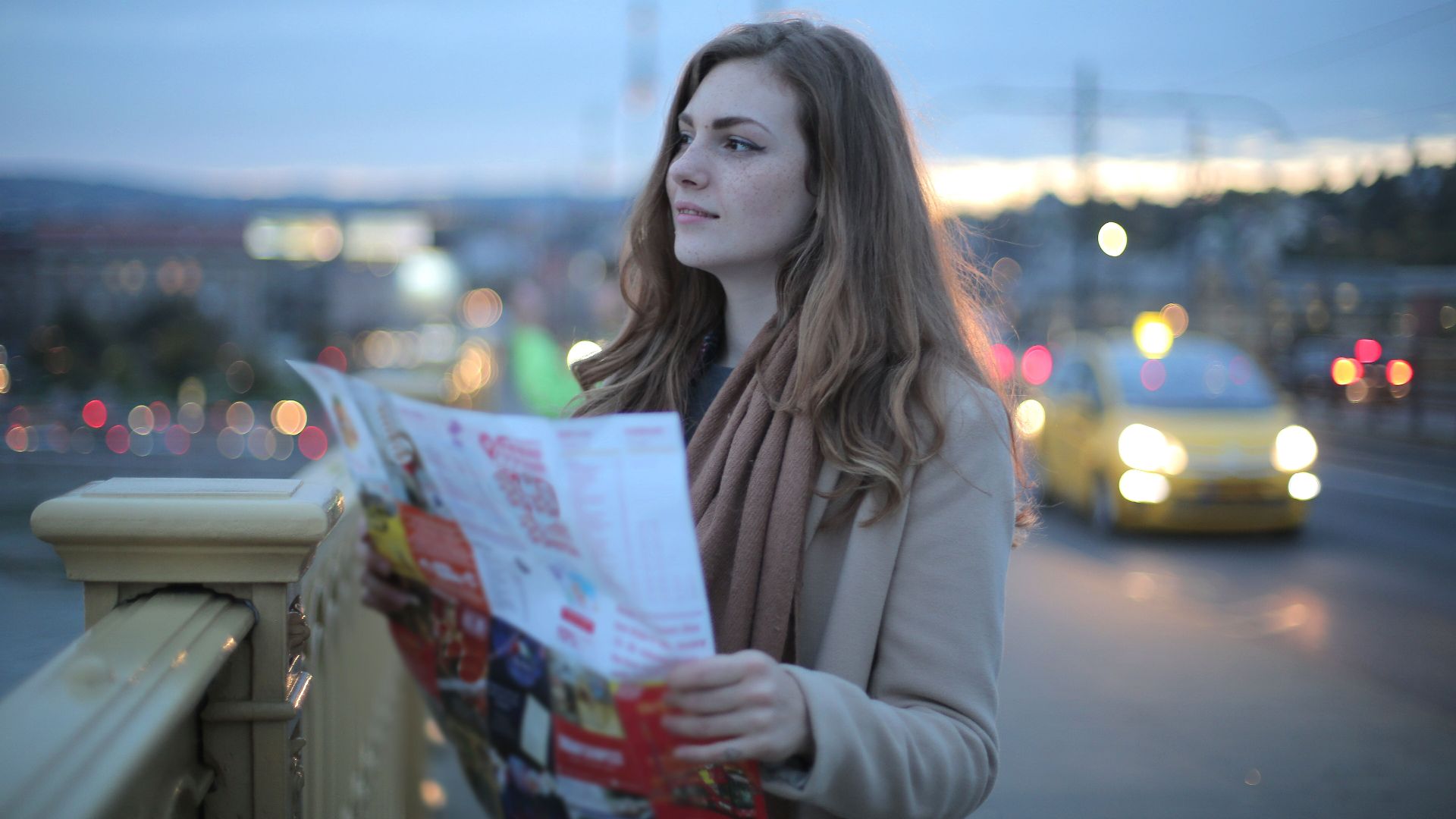Female traveler in warm clothes standing on street with map and looking away at dusk