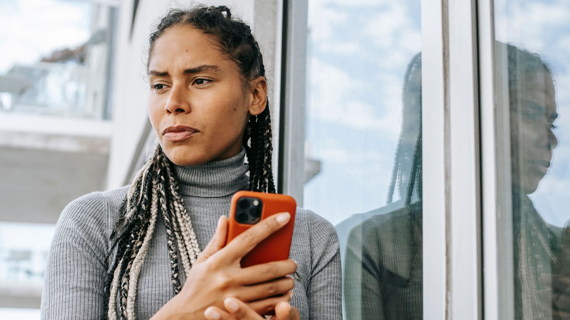 Low angle of concerned ethnic female with smartphone in stressful situation near glass wall