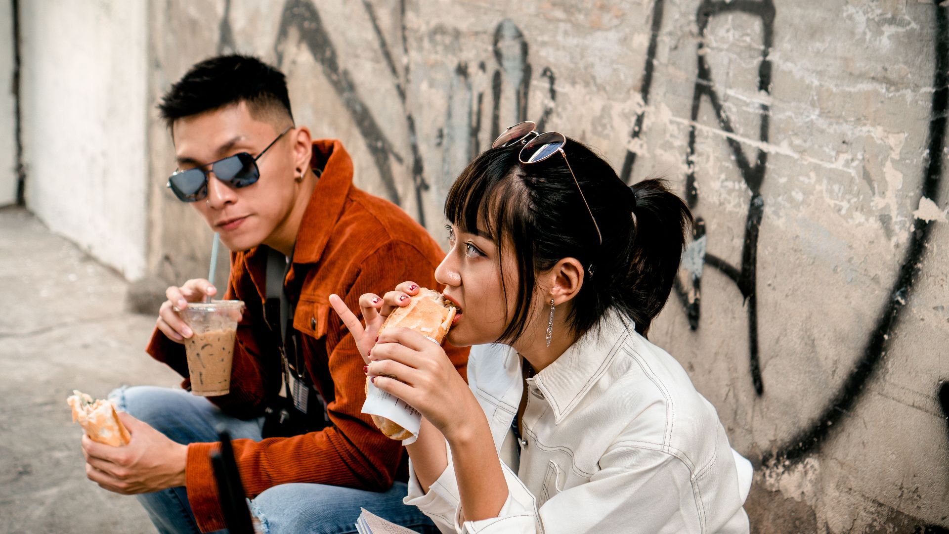Young urban adults in casual wear enjoying street food outdoors against a graffiti wall.