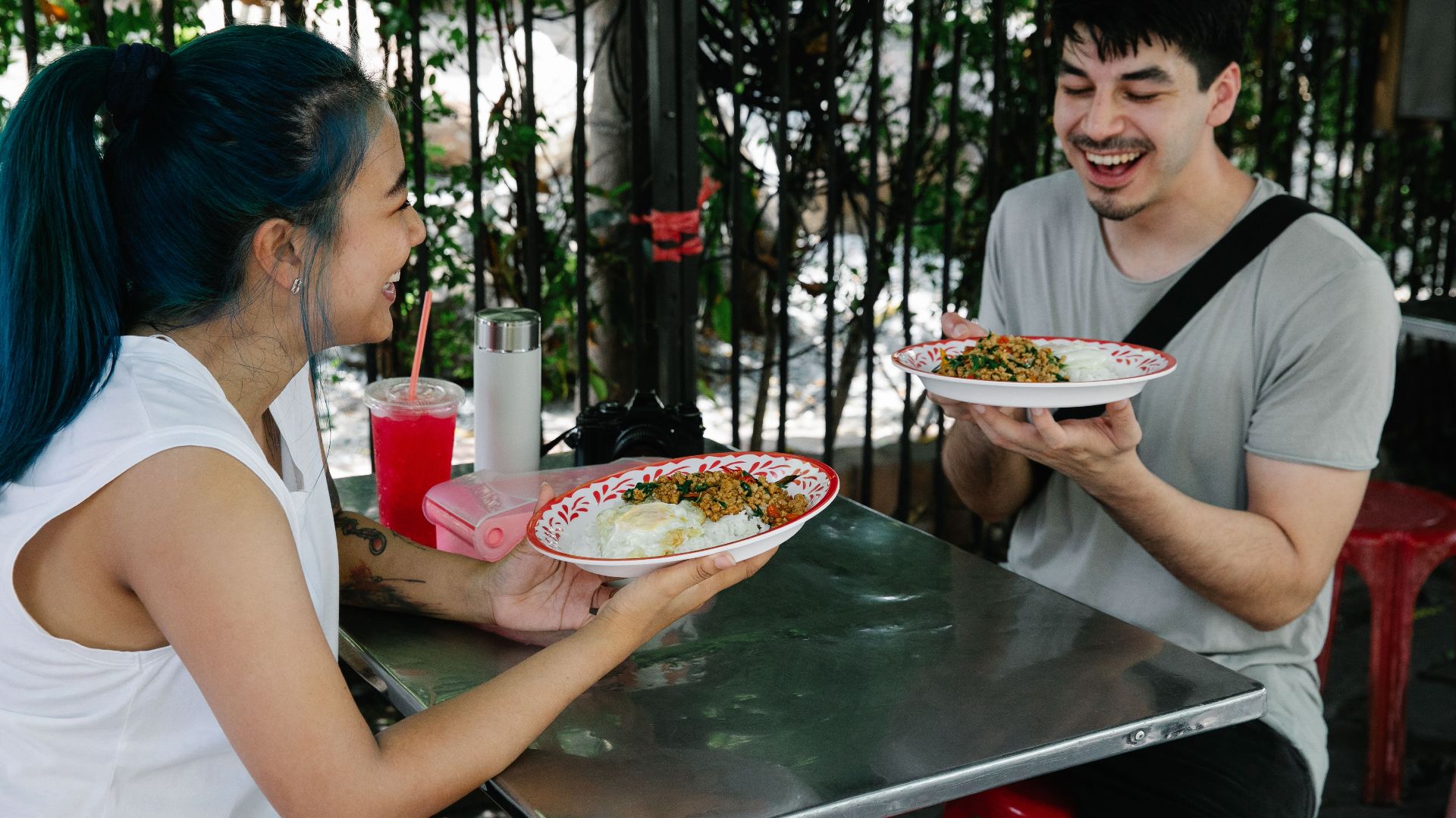 Happy multiracial couple sitting at table with plates of delicious food while having lunch together in outdoor cafeteria on summer day