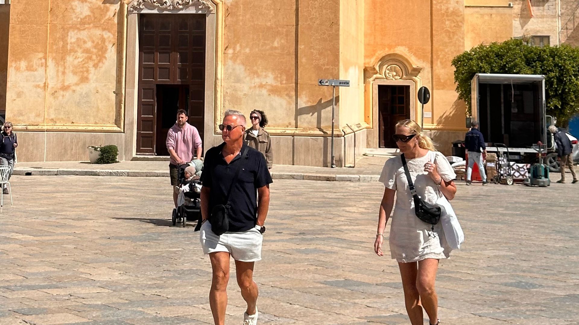Tourists walk past an old church with a distinctive bell tower under a clear blue sky.