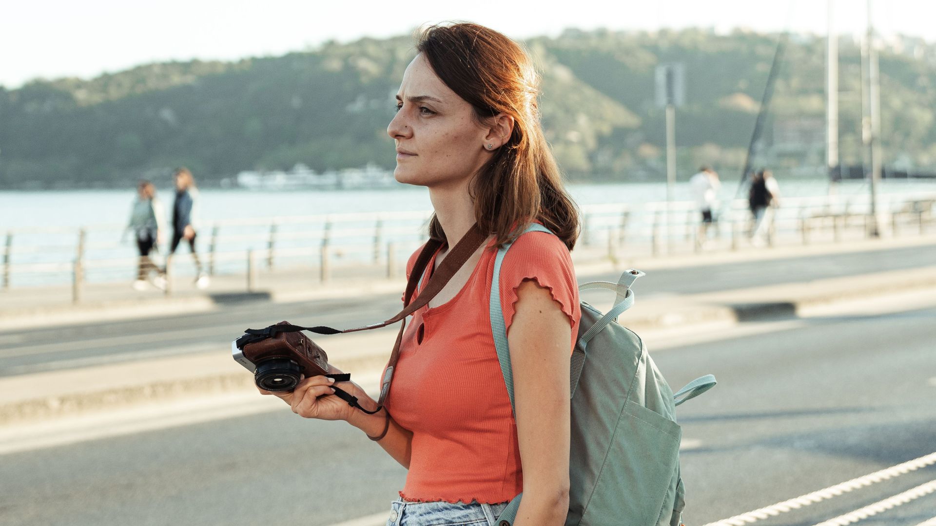 Woman standing by the waterfront with a camera and backpack on a sunny day.