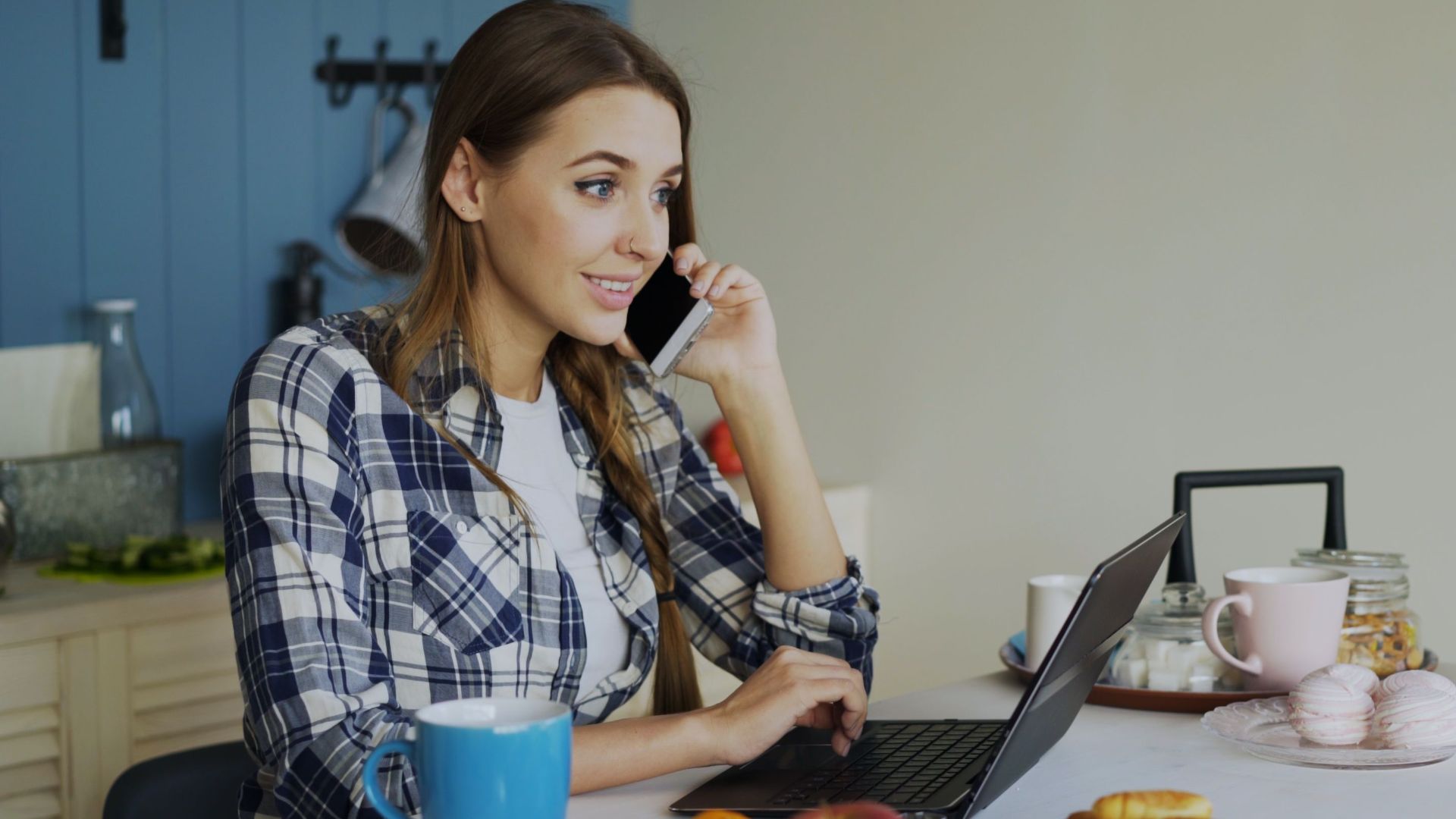 Young woman multitasking on phone and laptop in a cozy kitchen setting.