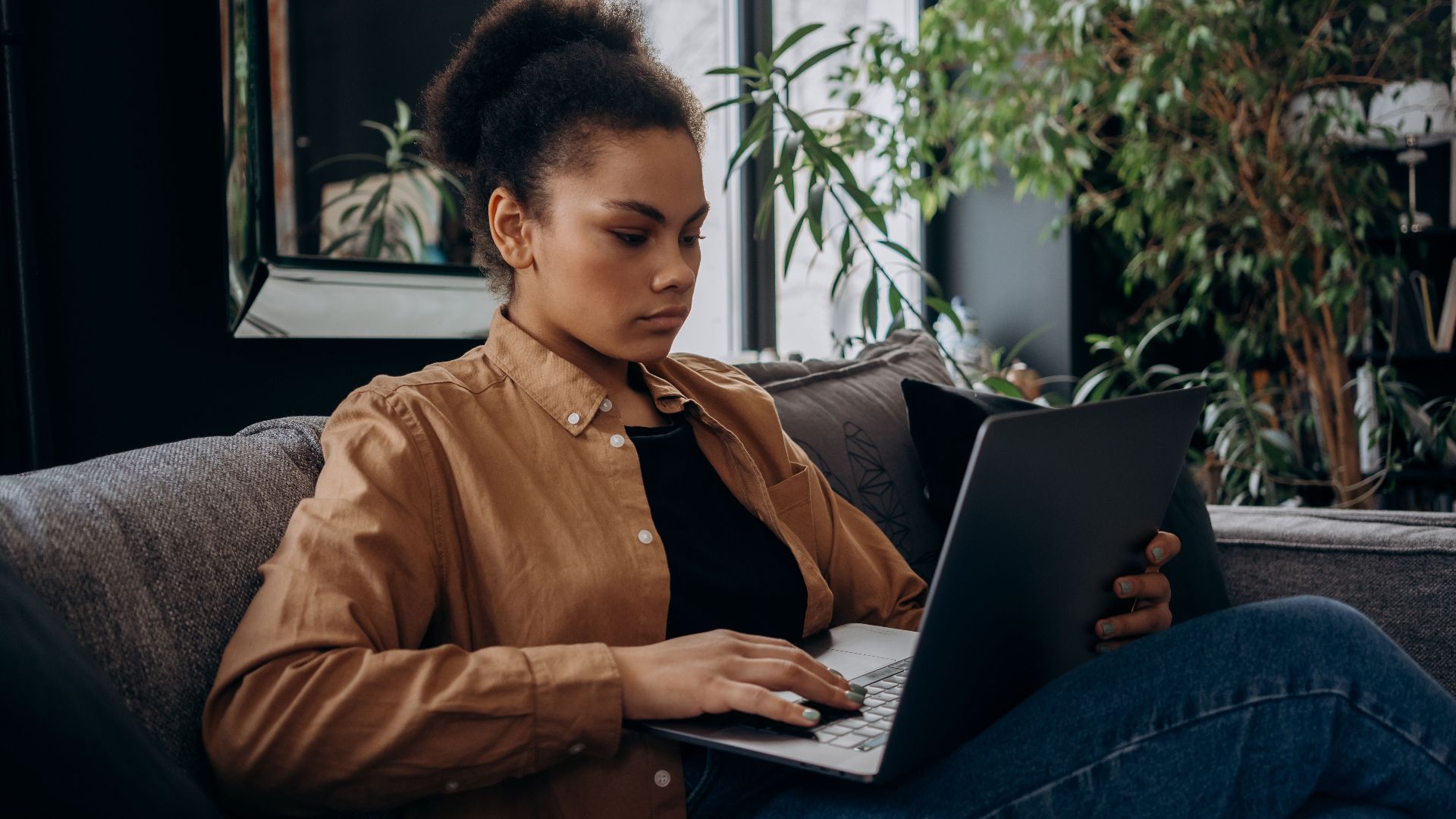 Focused young woman using a laptop on the couch, working remotely indoors.