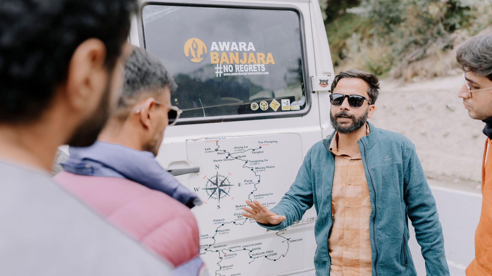 A guide in a blue jacket explains a travel route to three adults near a vehicle