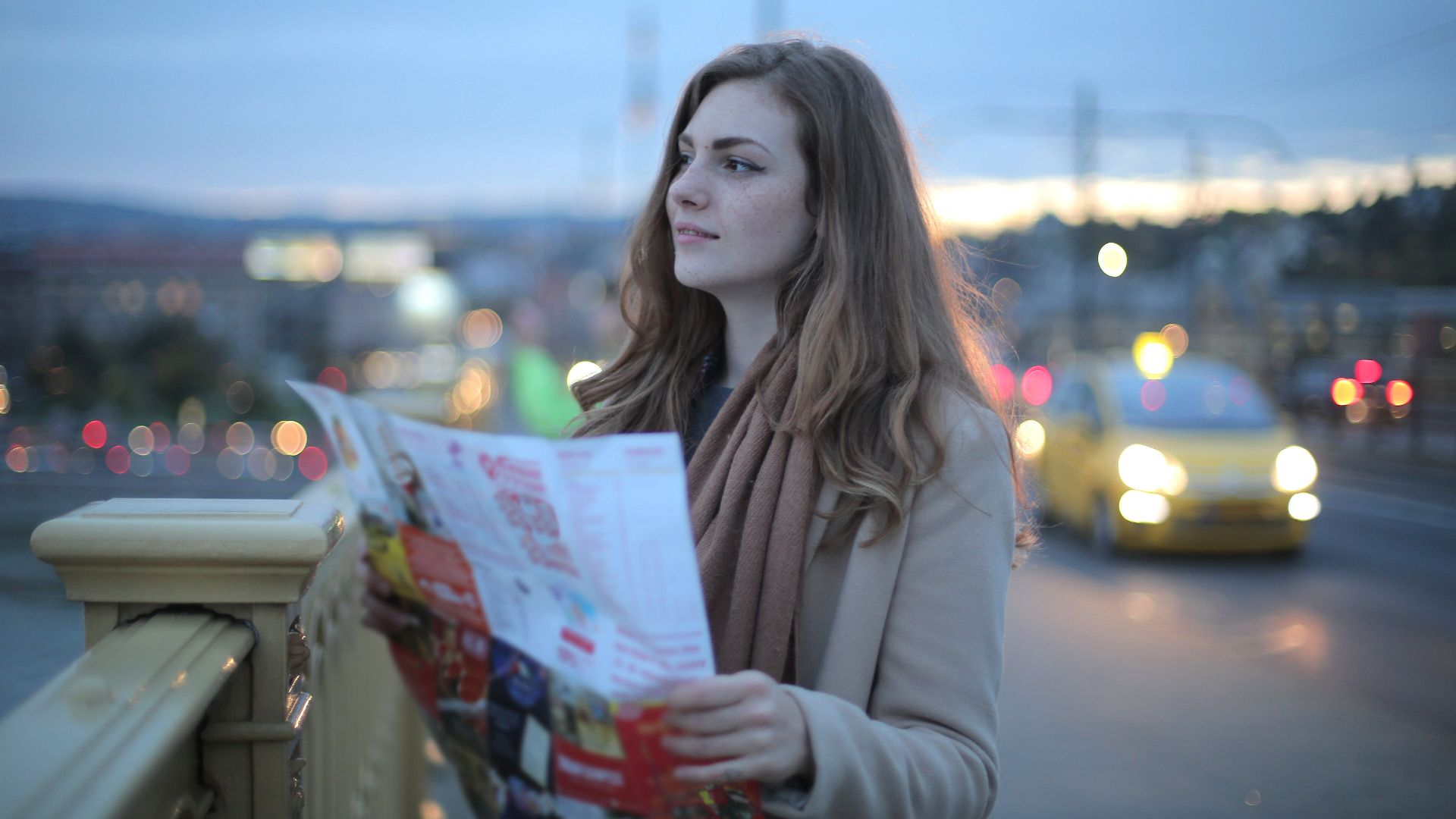 Female traveler in warm clothes standing on street with map and looking away at dusk