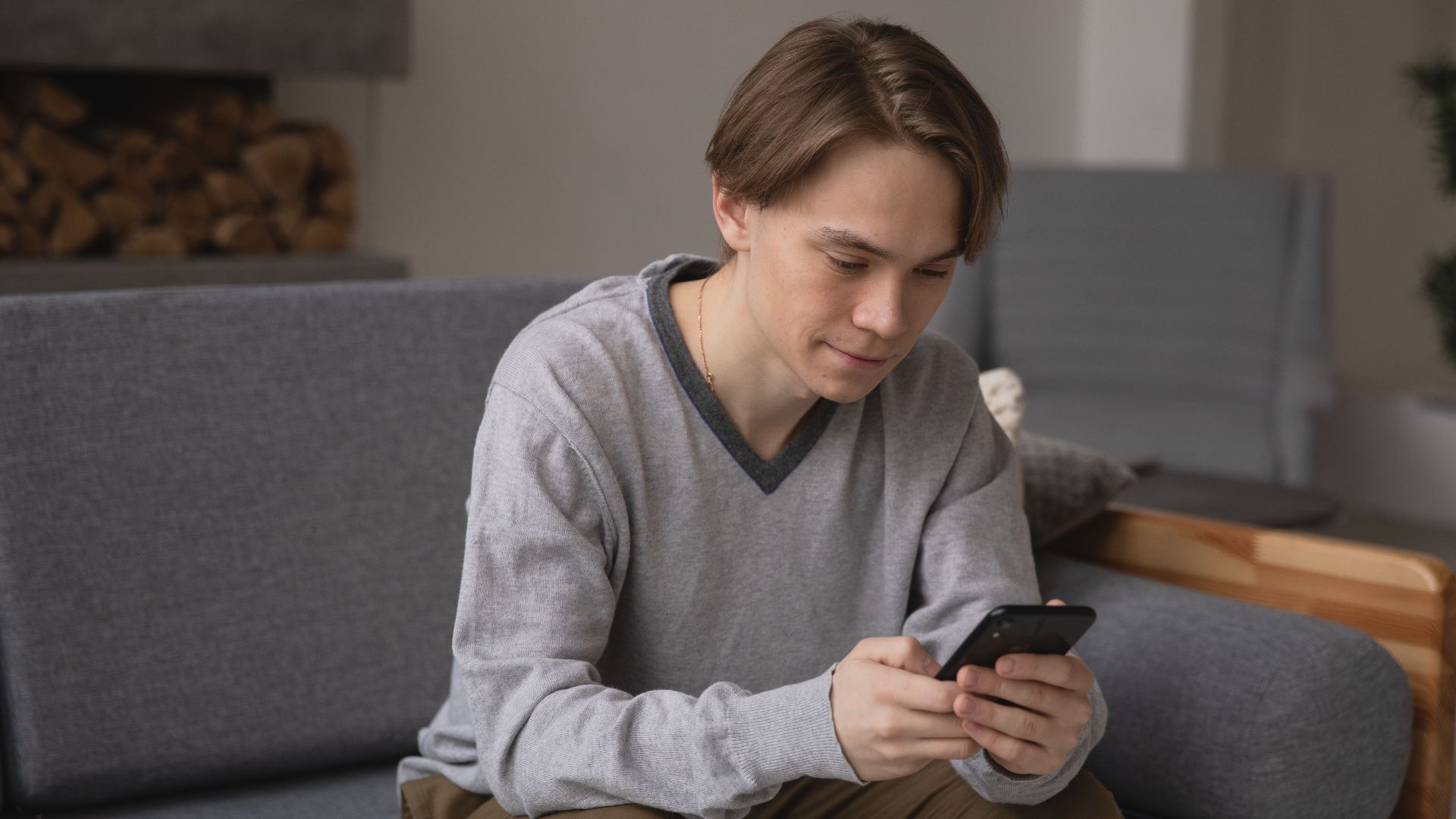 A young man comfortably sitting on a gray couch using his smartphone indoors.