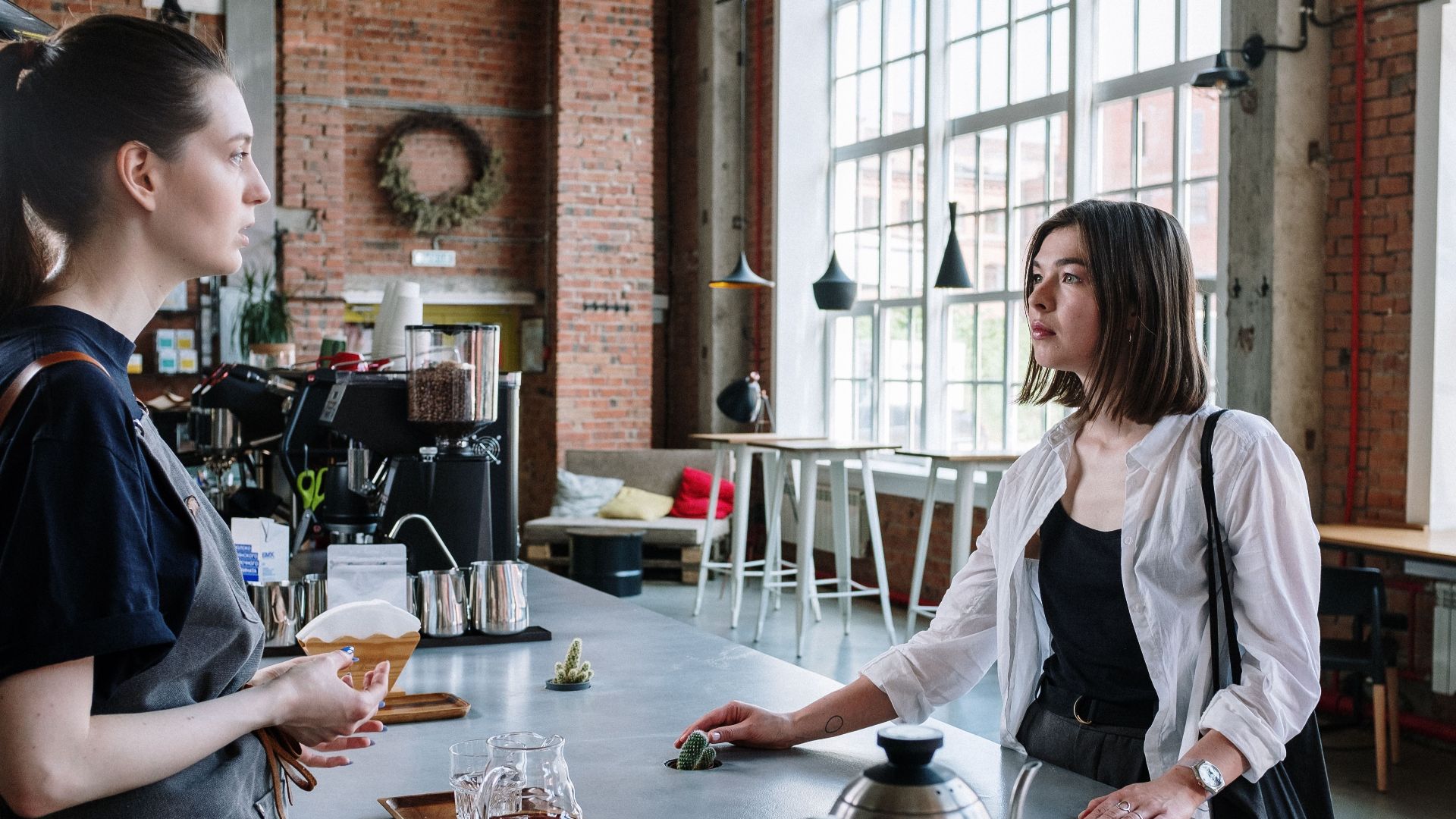 A barista and customer engage in a conversation at a stylish café counter with exposed brick walls.