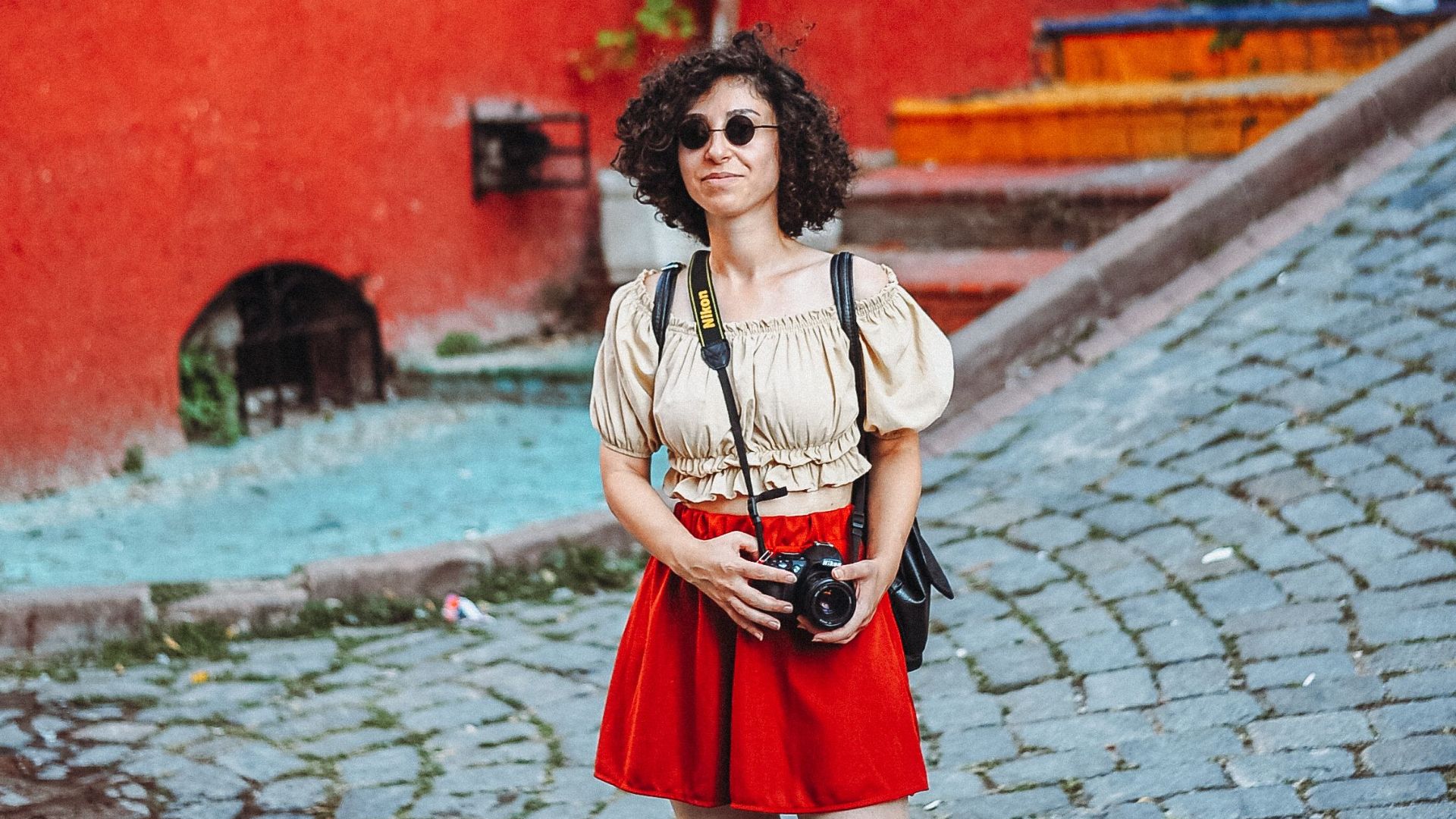 Woman with camera exploring vibrant old town, wearing red skirt and sunglasses, capturing travel moments.