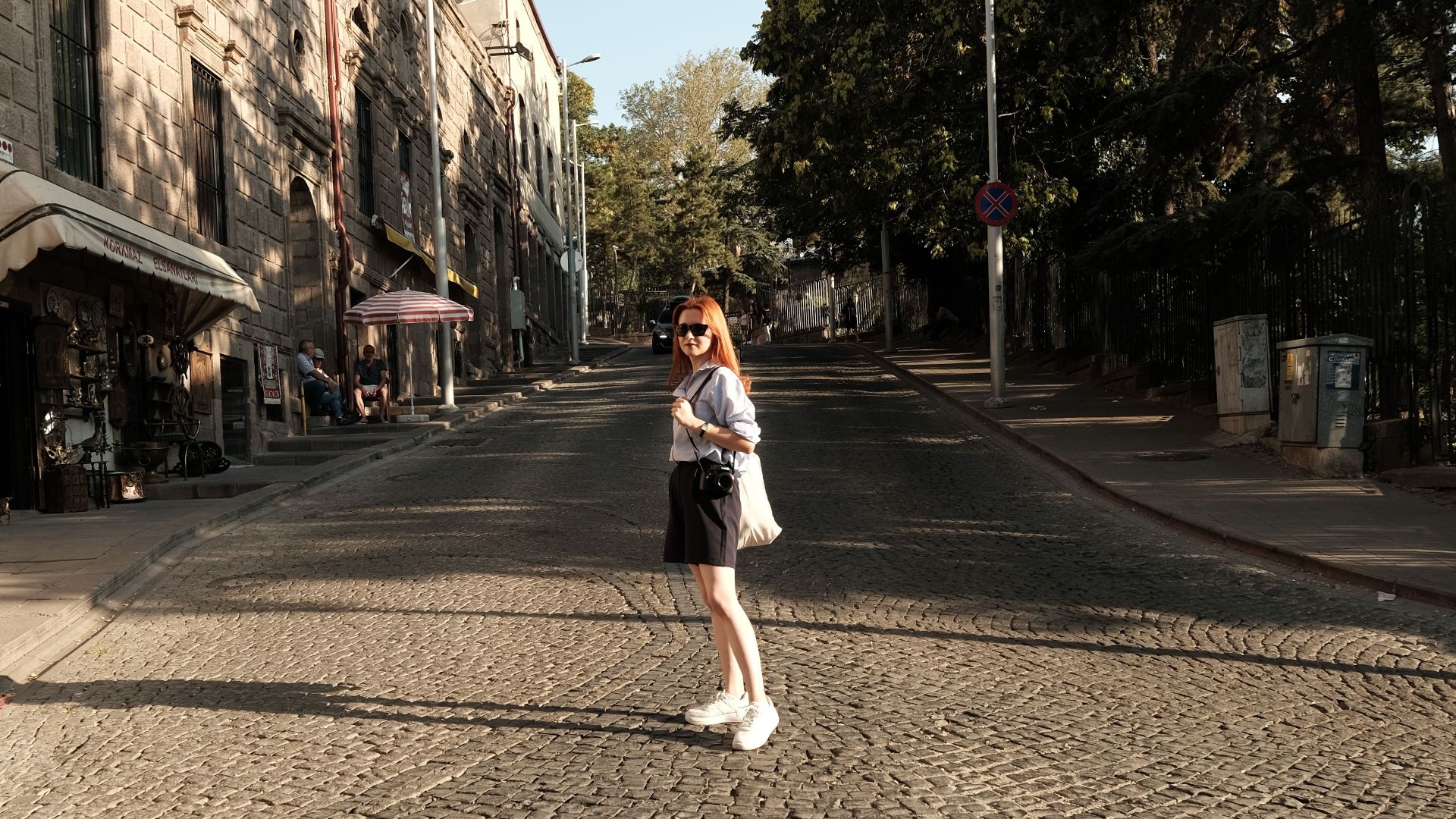 Woman in sunglasses standing on a cobblestone street lined with buildings and trees.