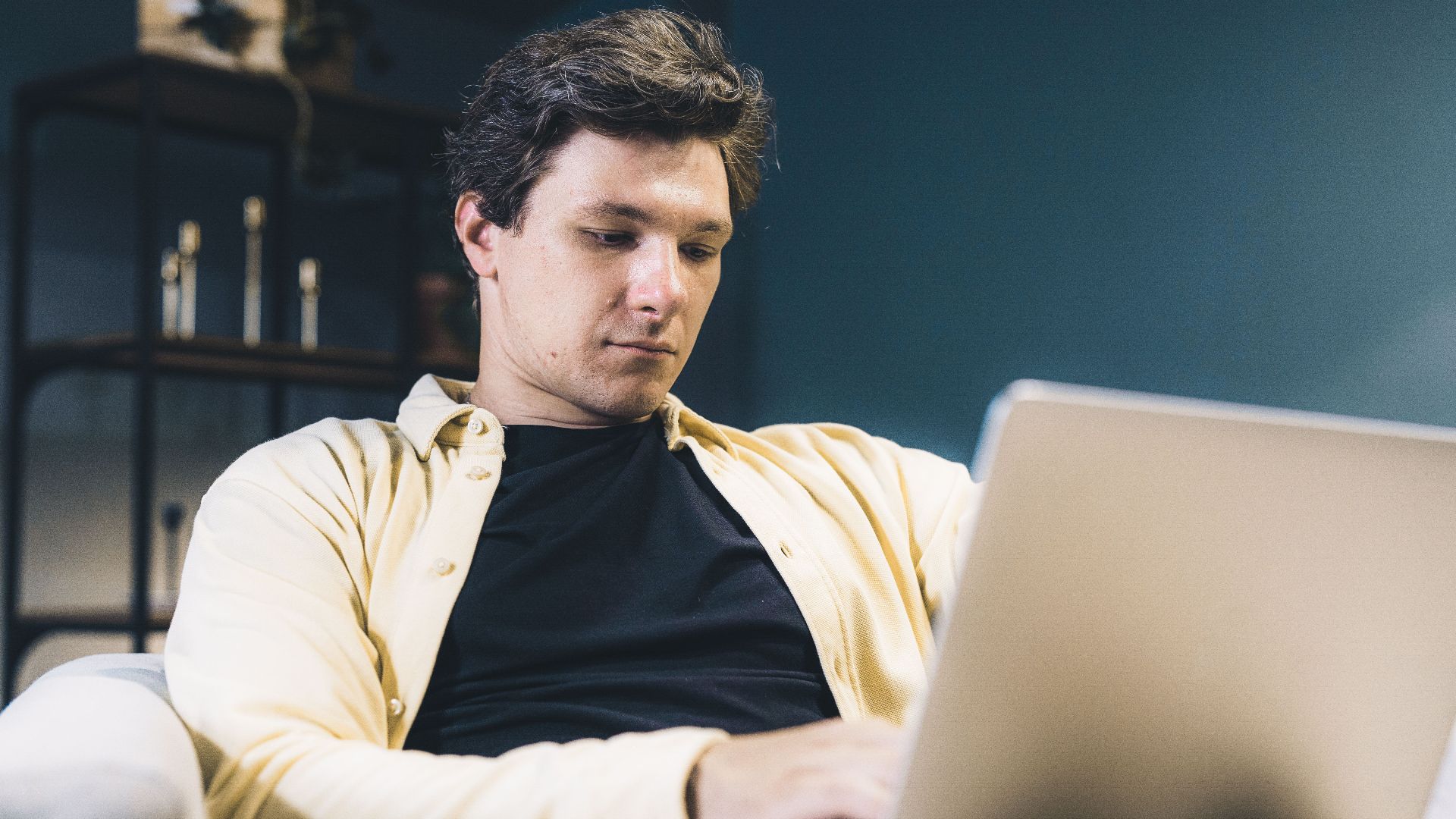 Focused young man sitting comfortably using a laptop at home, engaging in remote work.