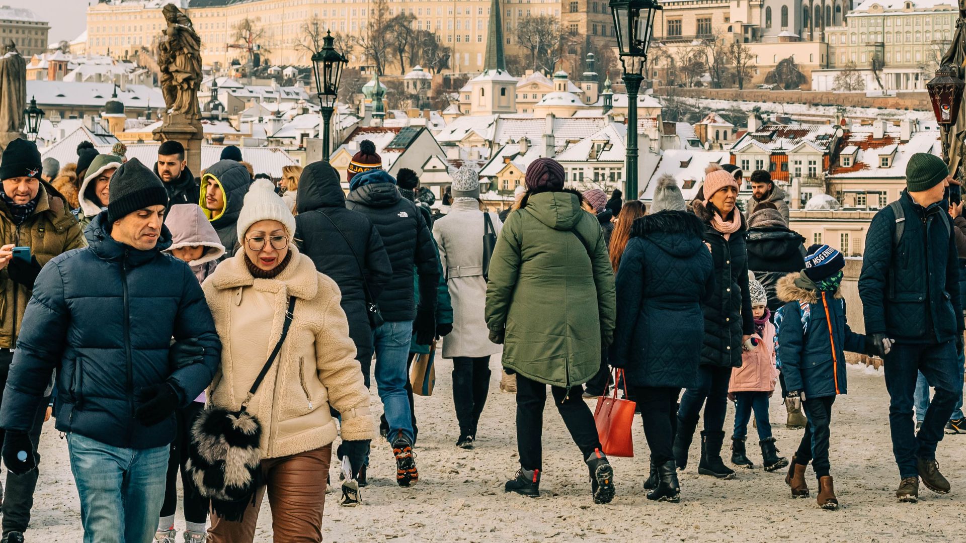 People walking on snow-covered Charles Bridge with Prague Castle in the background.