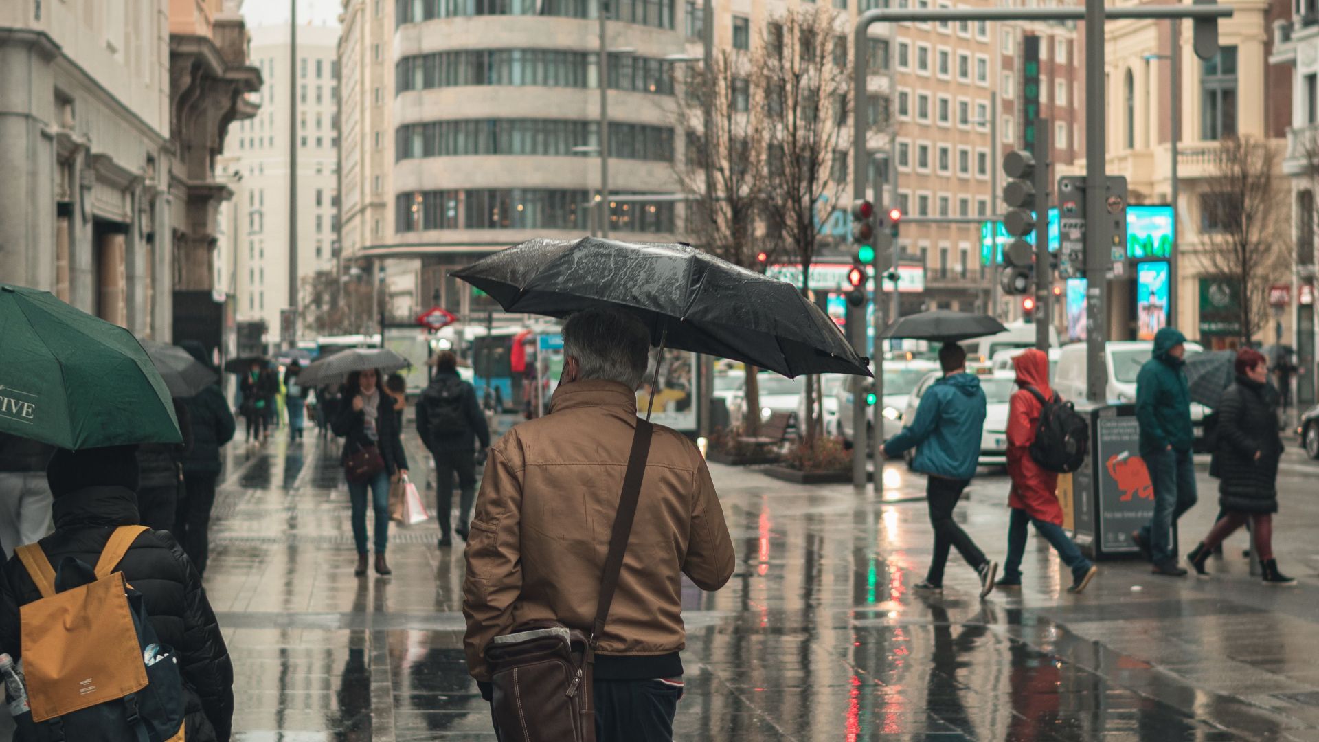 People with umbrellas walking on a wet street in Madrid, featuring iconic Schweppes building.