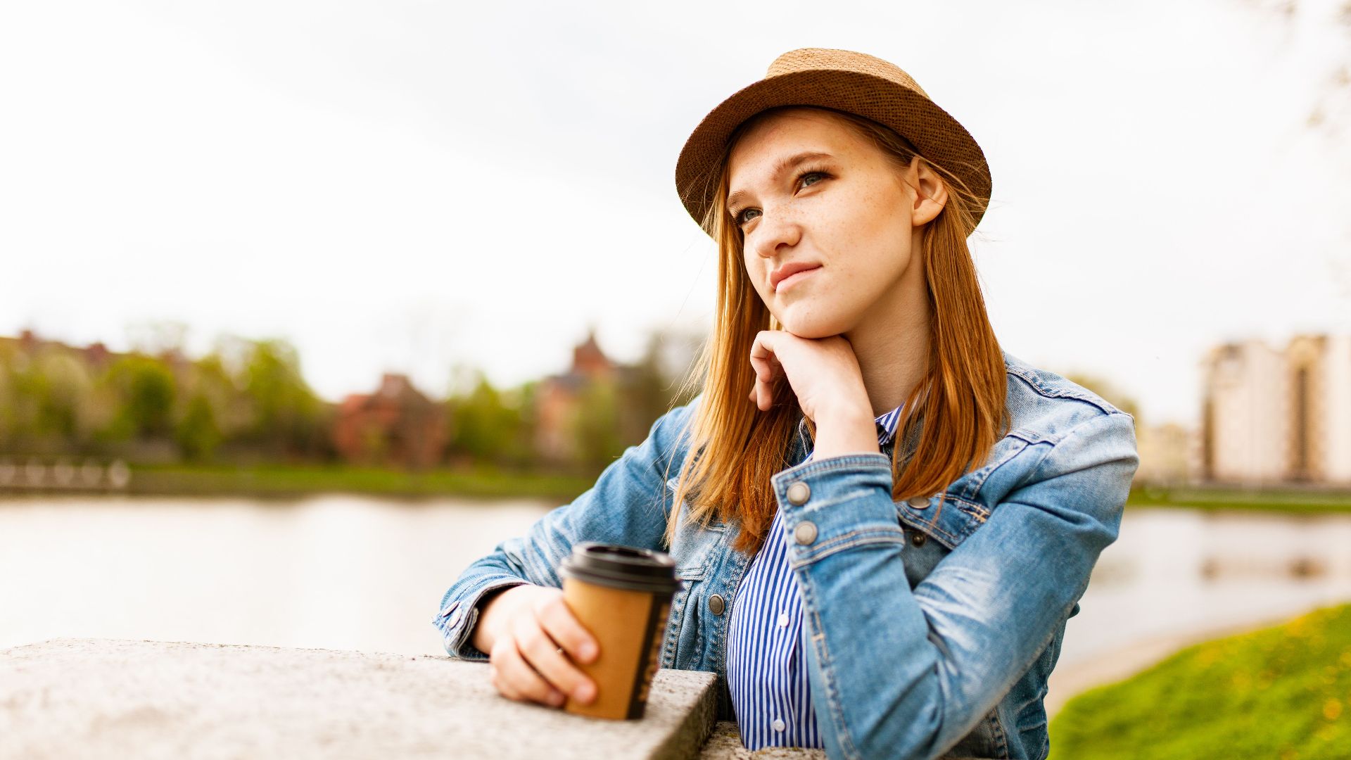 Woman in denim and hat enjoying coffee outdoors with a thoughtful expression by the lake.
