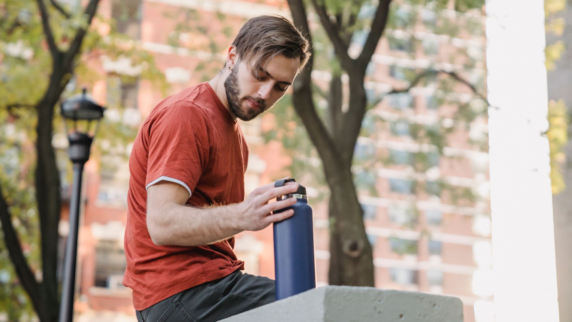 Low angle side view of young unshaven male athlete with bottle resting on fence in town