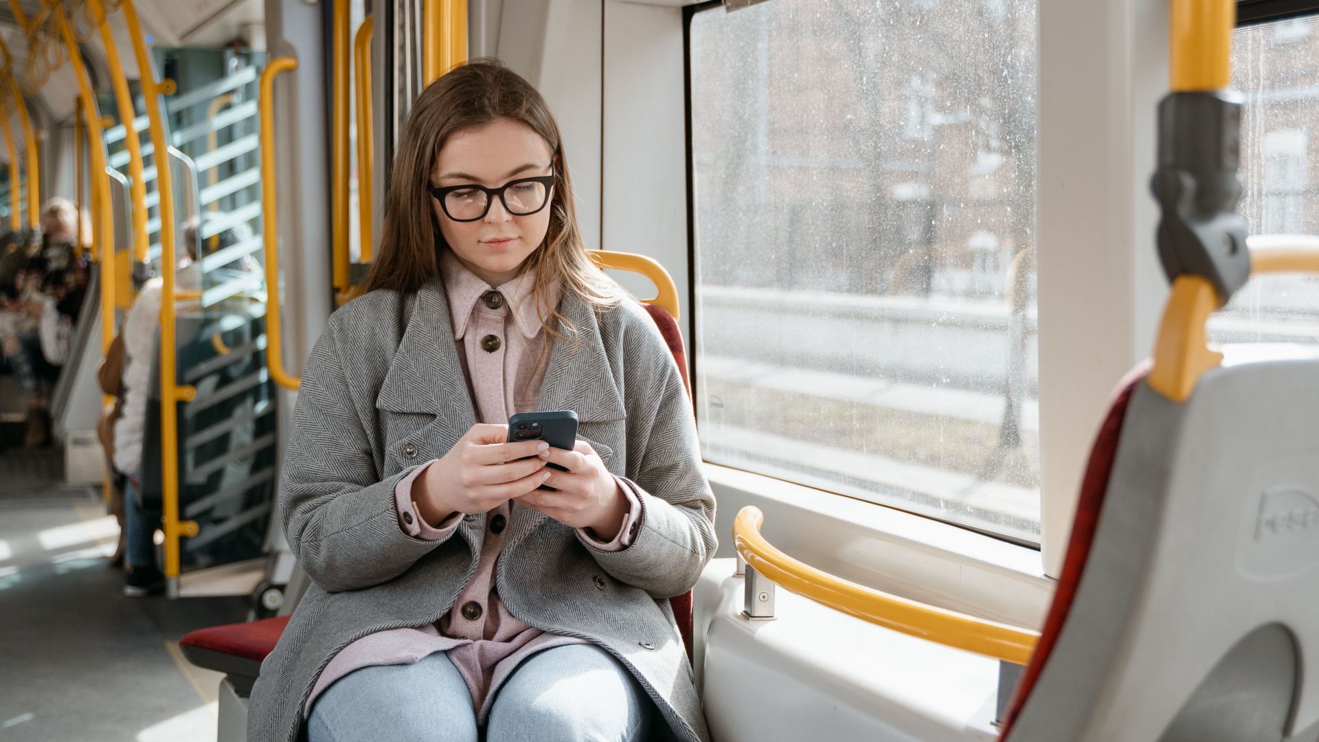 Caucasian woman using smartphone on a tram, wearing a gray coat in an urban setting.