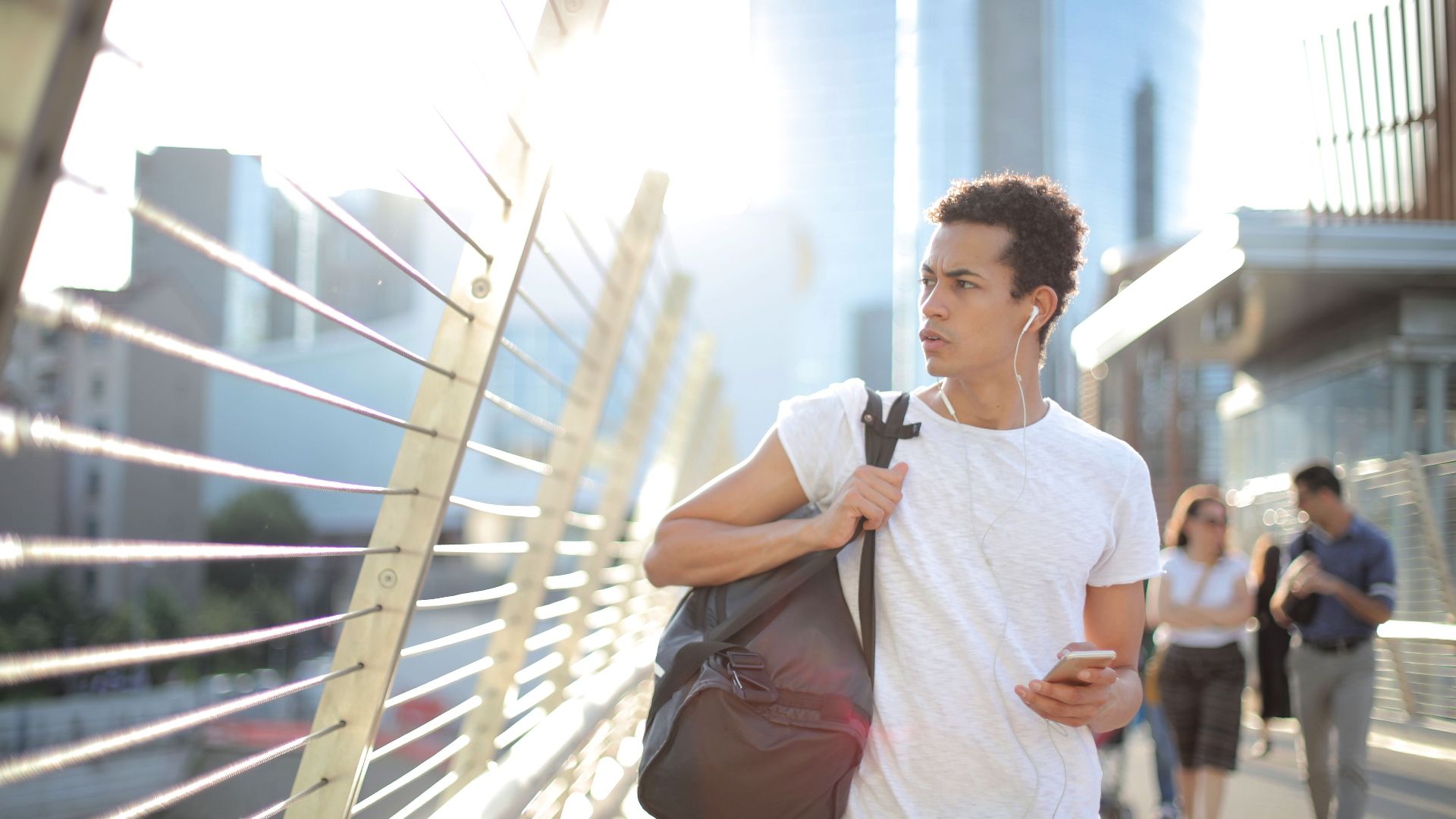 Concentrated guy with smartphone and sportive bag in hands wearing earphones walking on bridge while listening to music