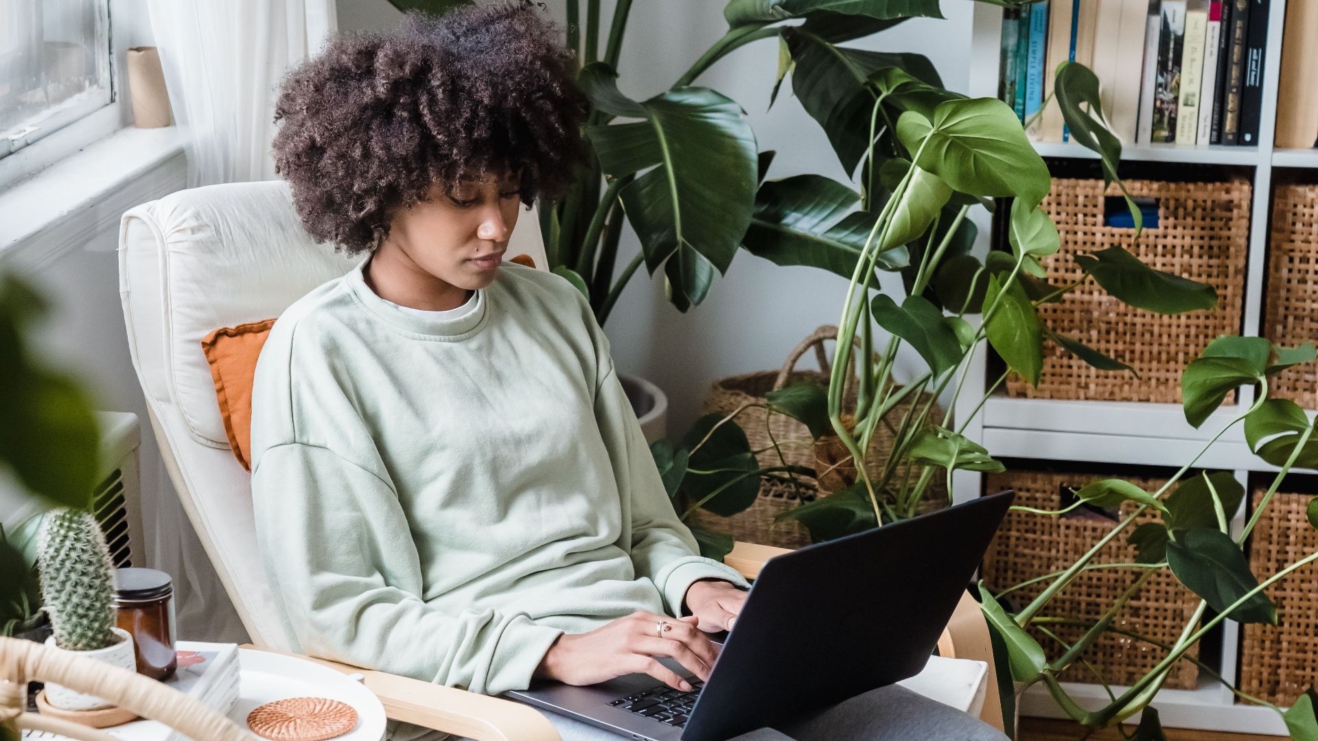 Woman in comfortable clothing using a laptop in a plant-filled cozy home setting.