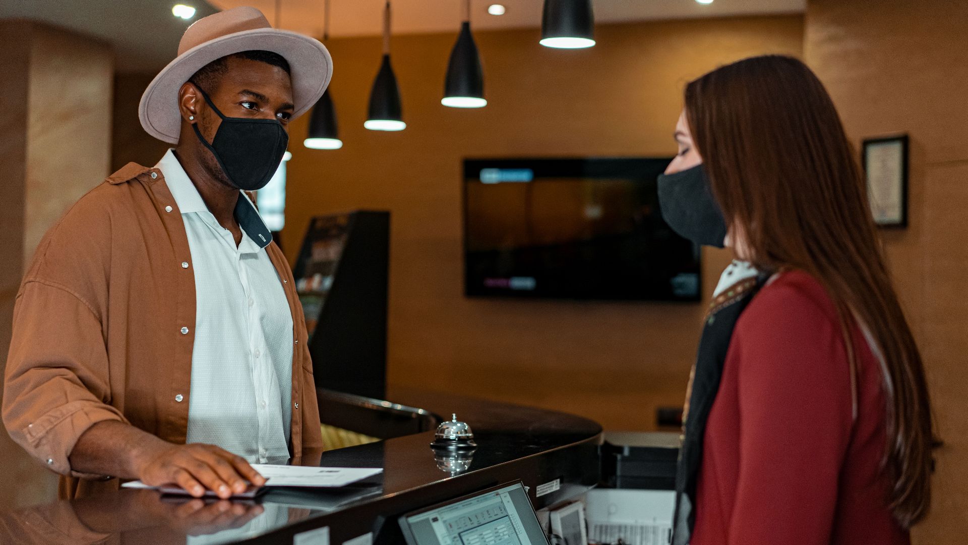 A man and woman in face masks checking in at a hotel reception, emphasizing safety protocols.