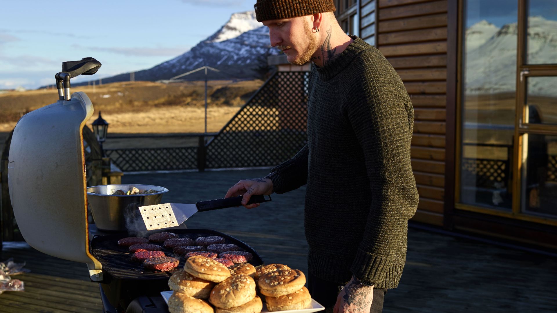 Side view of young man in casual warm clothes roasting meat for burger on veranda of country restaurant in valley among snowy hills