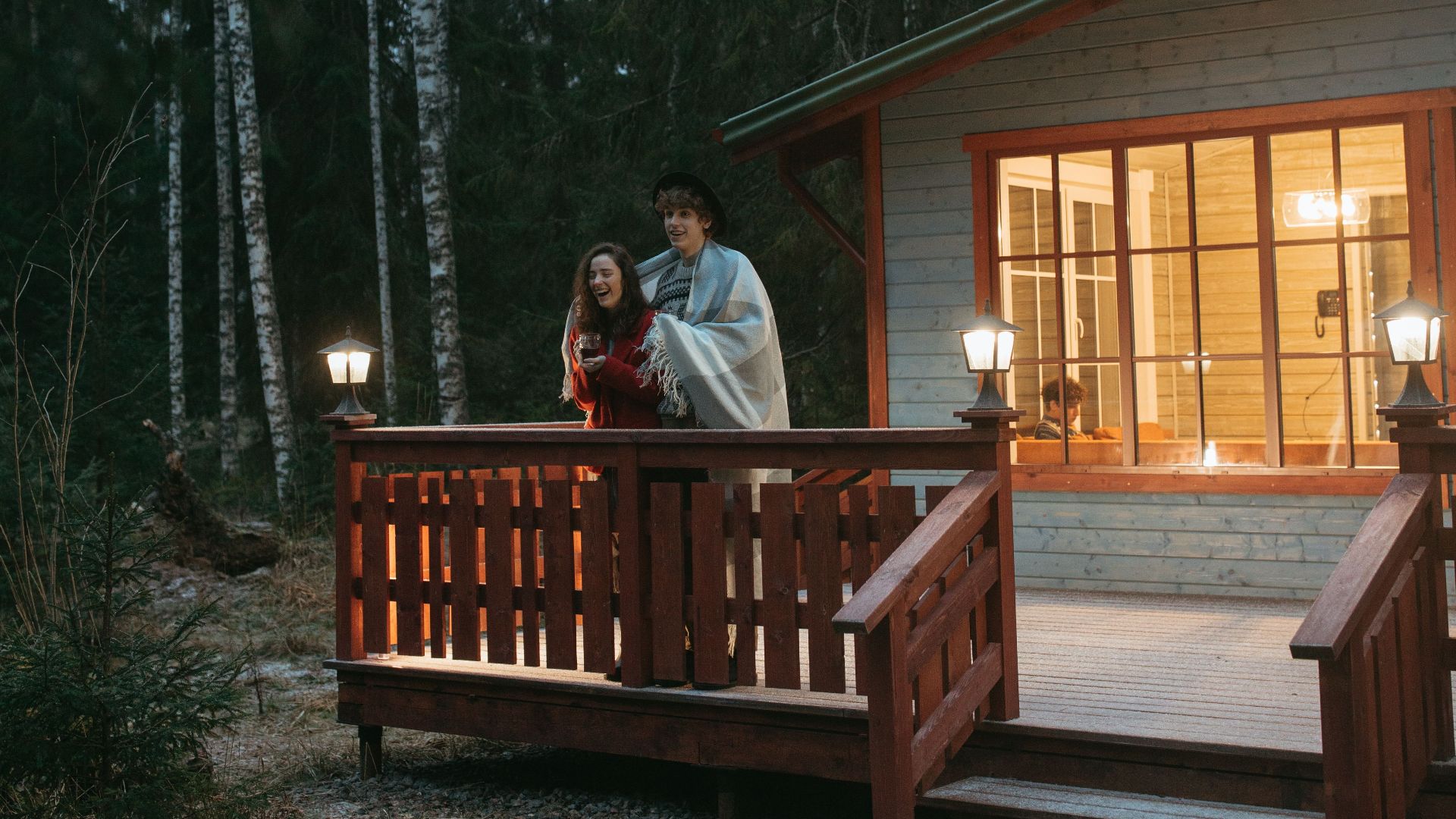 A couple wrapped in a blanket enjoys a winter evening on a forest cabin balcony.