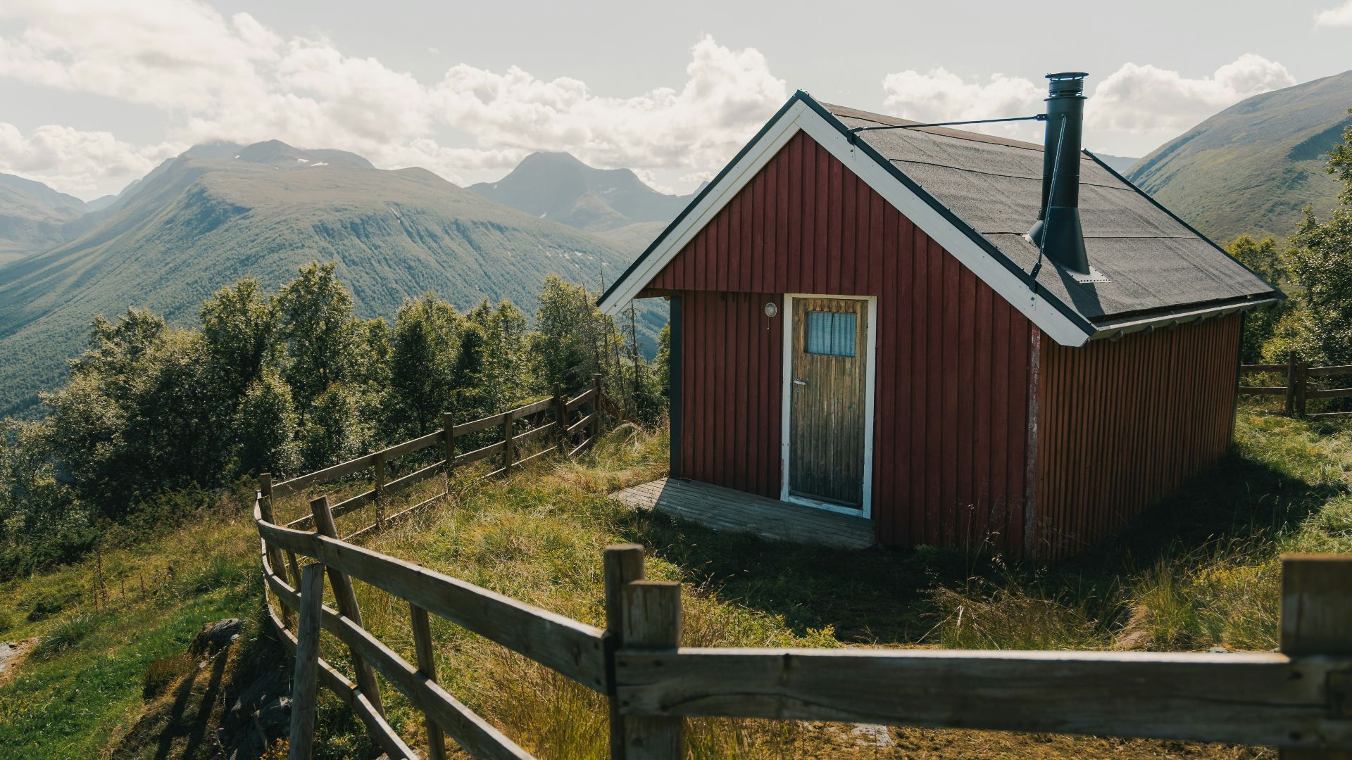 Red cabin on a hill with mountains and fence.