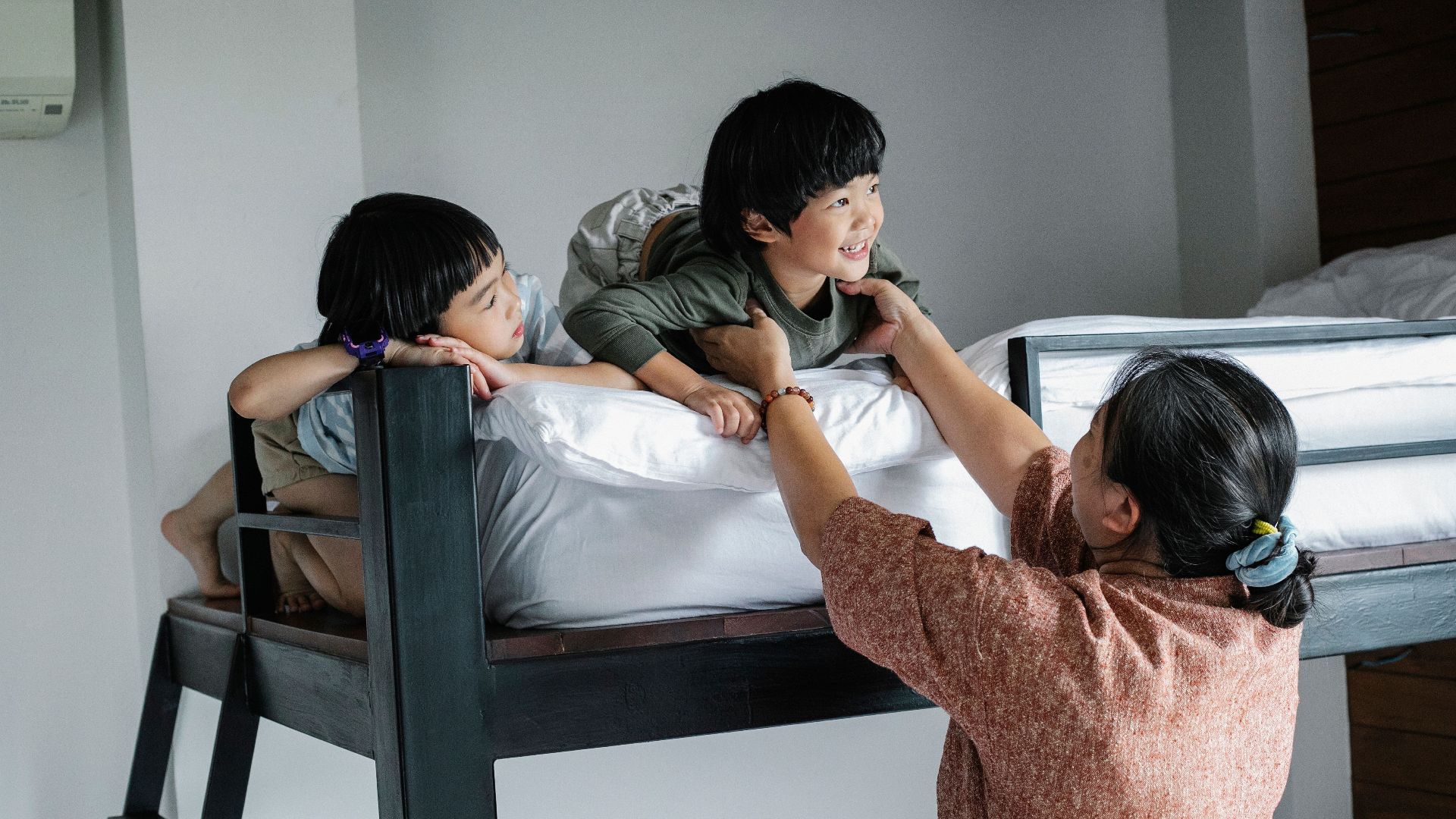 Back view of grandmother standing near bunk bed and reaching arms to Asian boy lying near girl in bedroom in morning
